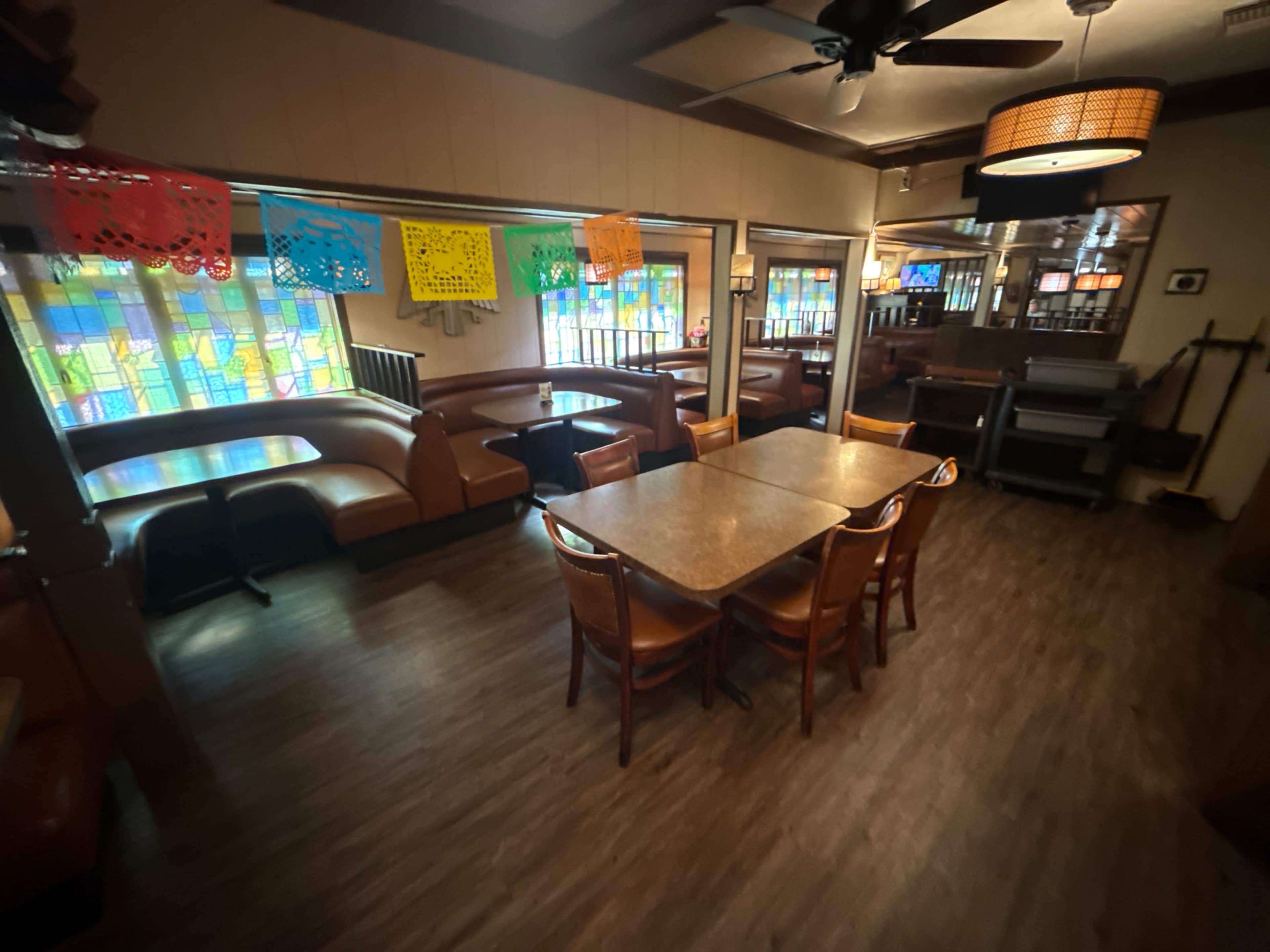 The image shows a cozy restaurant interior featuring wooden tables and chairs, colorful paper decorations, and booths along the walls with large windows allowing natural light to enter.