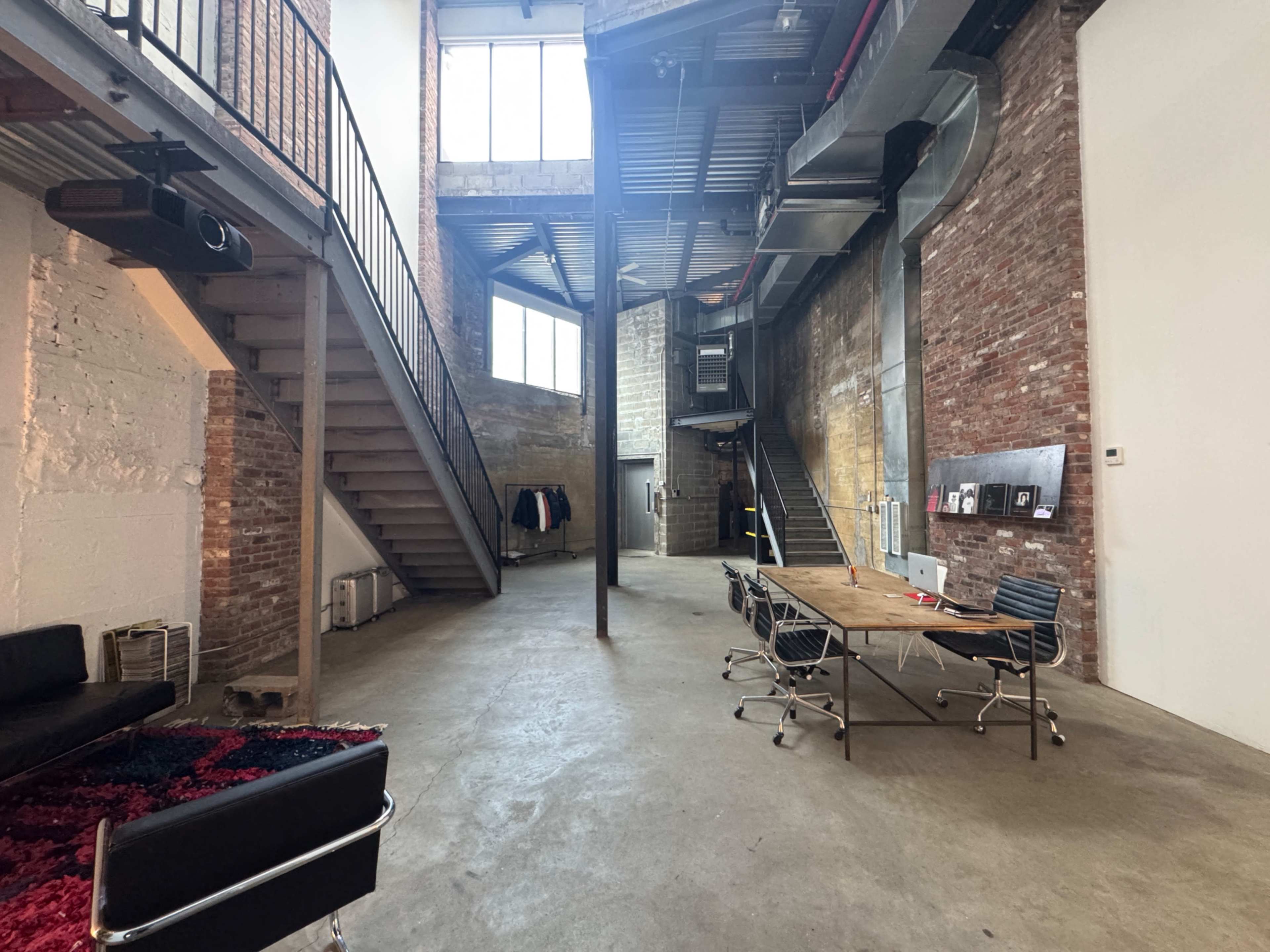 The image shows a spacious industrial-style room with exposed brick walls, large windows, a staircase, and a long wooden table surrounded by rolling chairs.