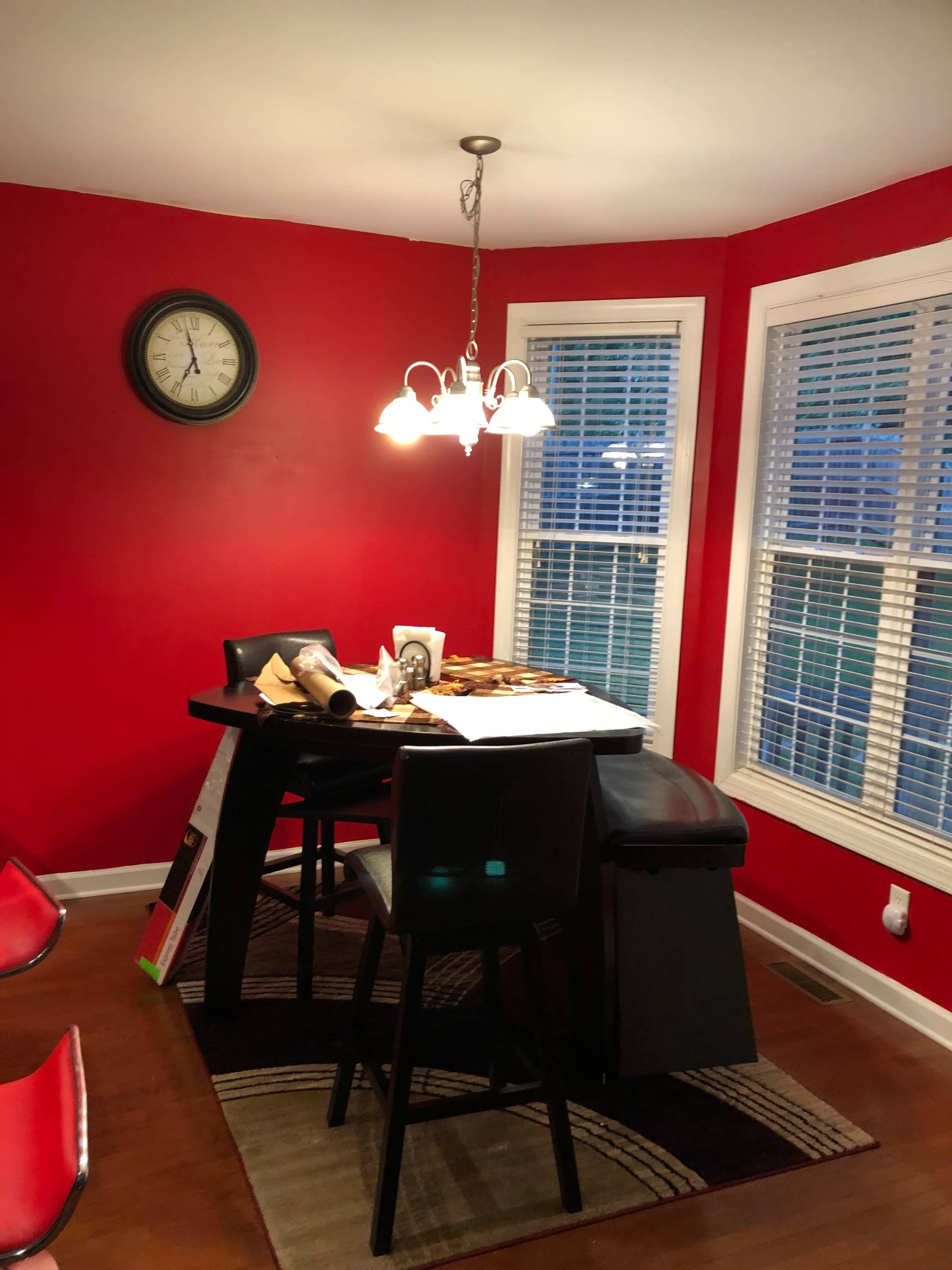 A dining area features a black table with chairs, a lamp overhead, and red walls, along with a clock and large windows with blinds.