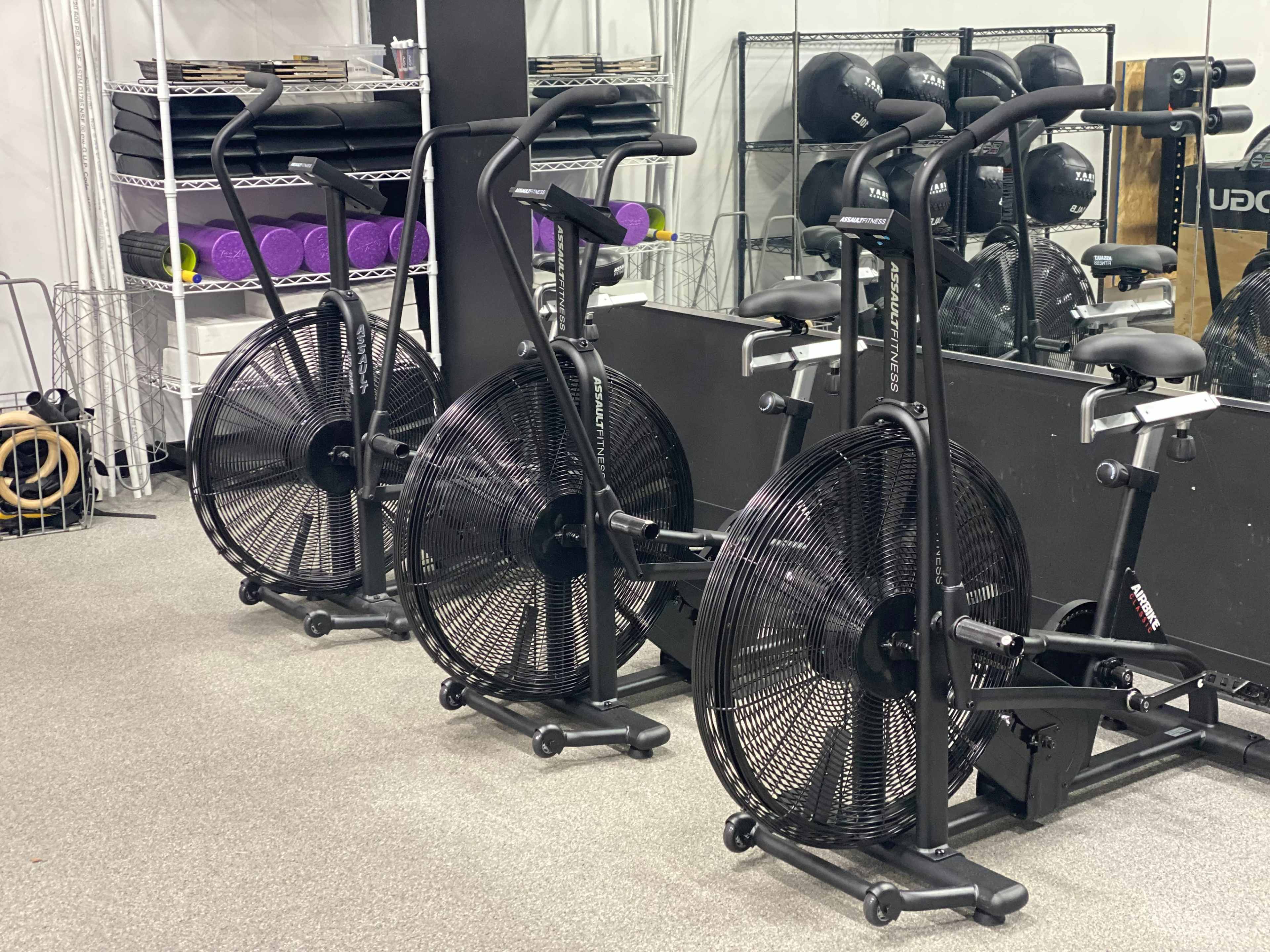 Three air bikes are lined up against a wall in a gym, with weights and exercise equipment visible in the background.
