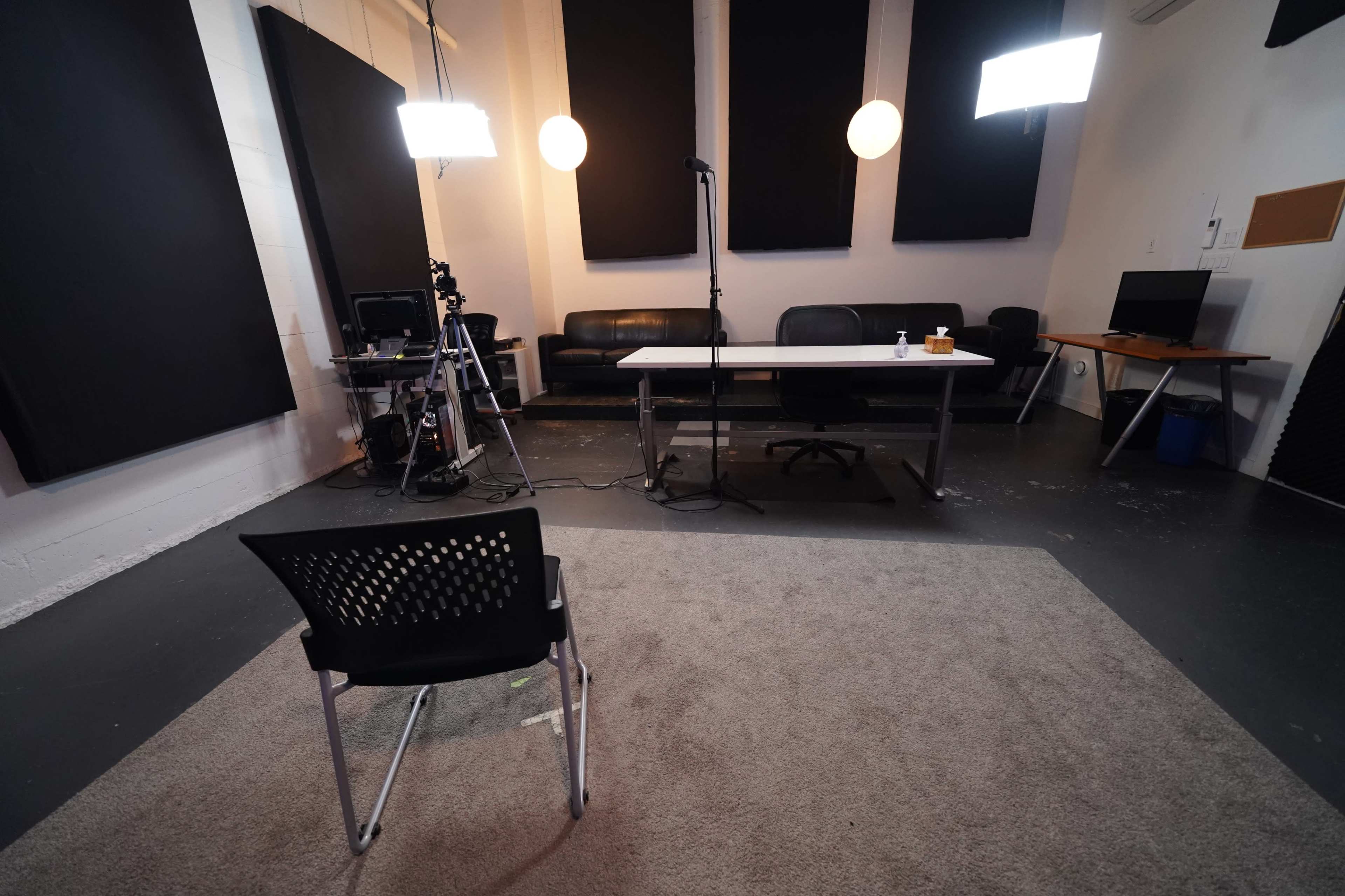 The image shows a recording studio with a microphone on a stand, a camera setup, and a single chair facing a table in a minimalistic, well-lit space.