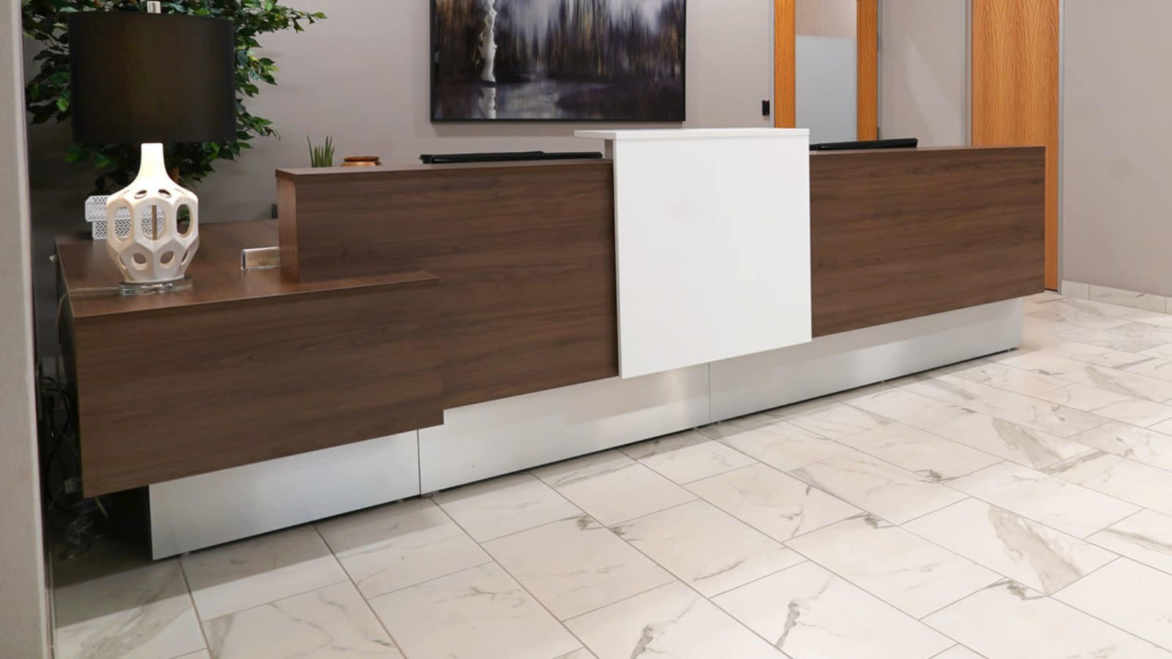 A modern reception desk made of wood and a white panel, positioned in an office with marble flooring and minimal decor.