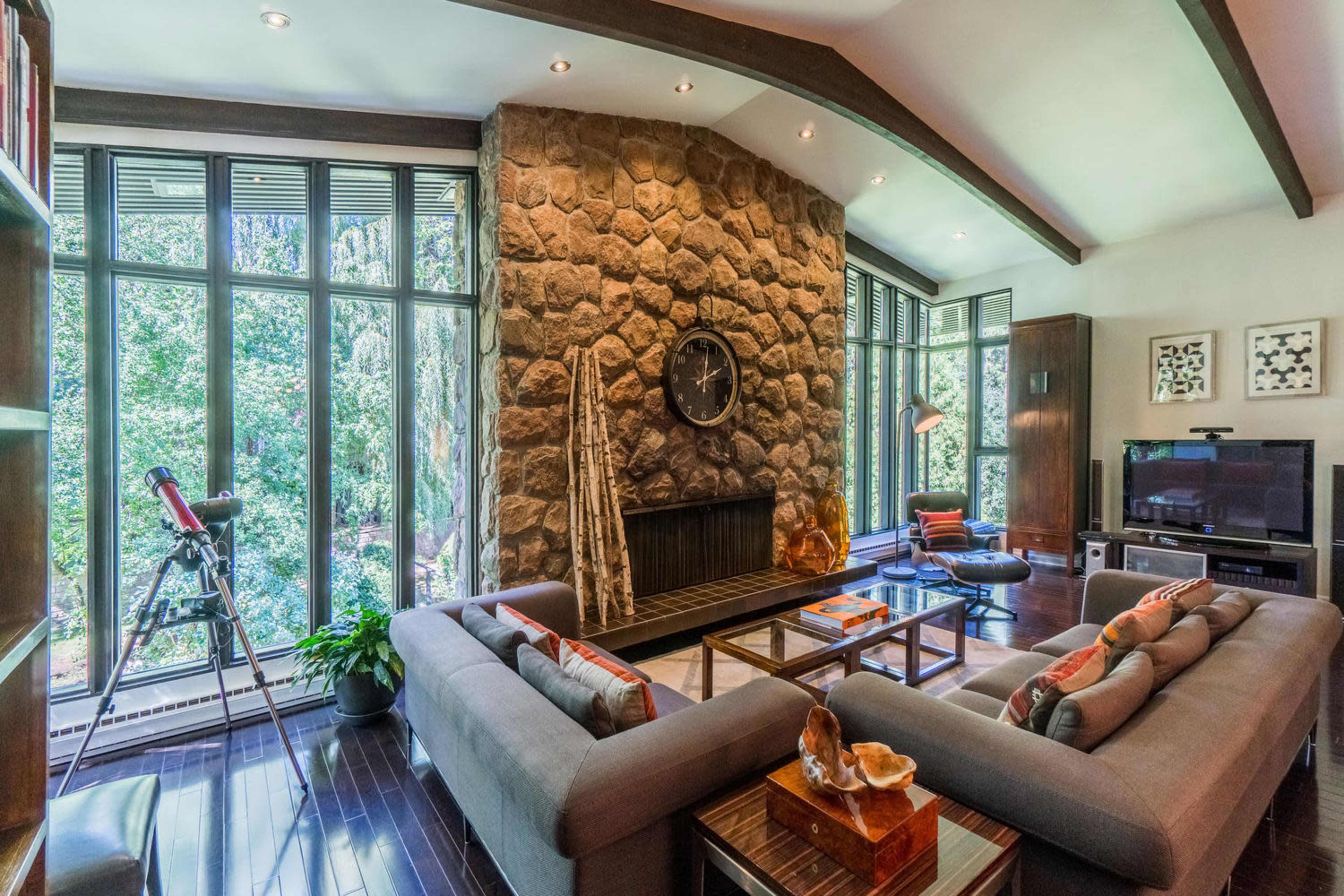 A modern living room featuring a large stone fireplace, large windows overlooking greenery, and contemporary furniture arranged around a coffee table.