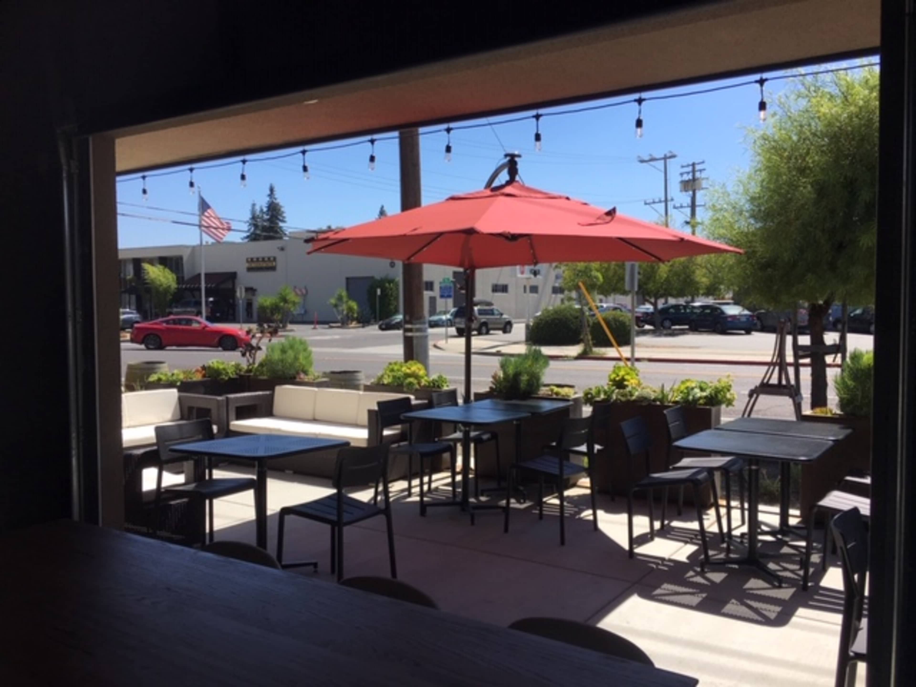 A view from inside a cafe shows a patio with tables and an umbrella, overlooking a street with a flag and parked cars.