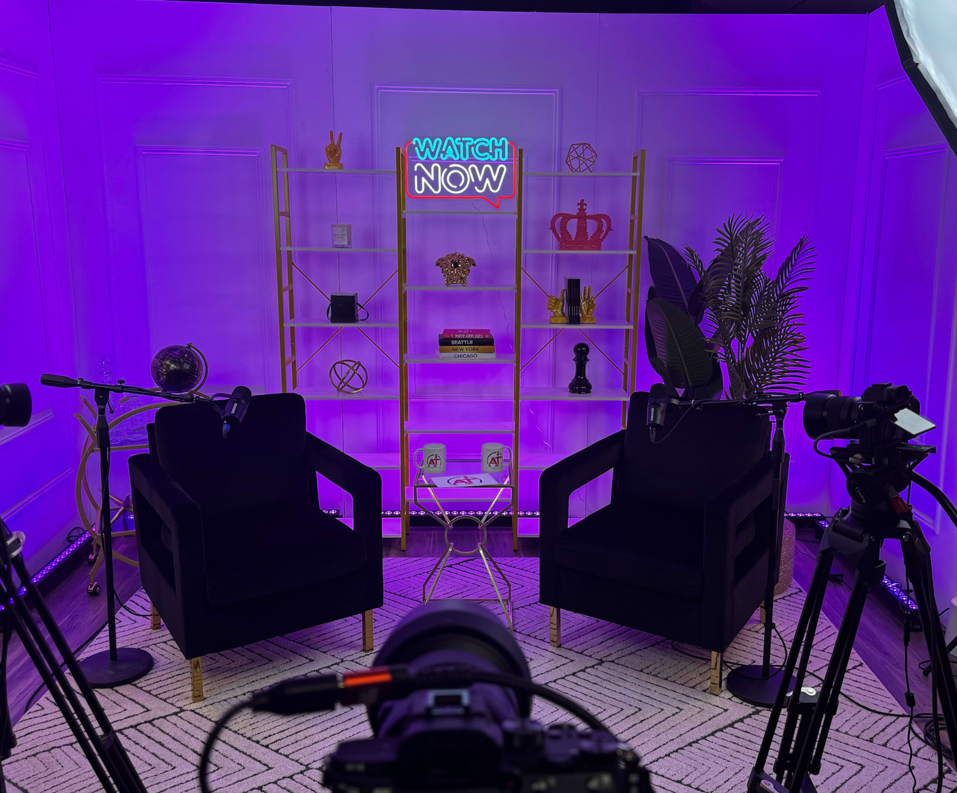 A recording studio setup with two black chairs facing a shelf decorated with various items, and a neon sign that reads "WATCH NOW," all under purple lighting.