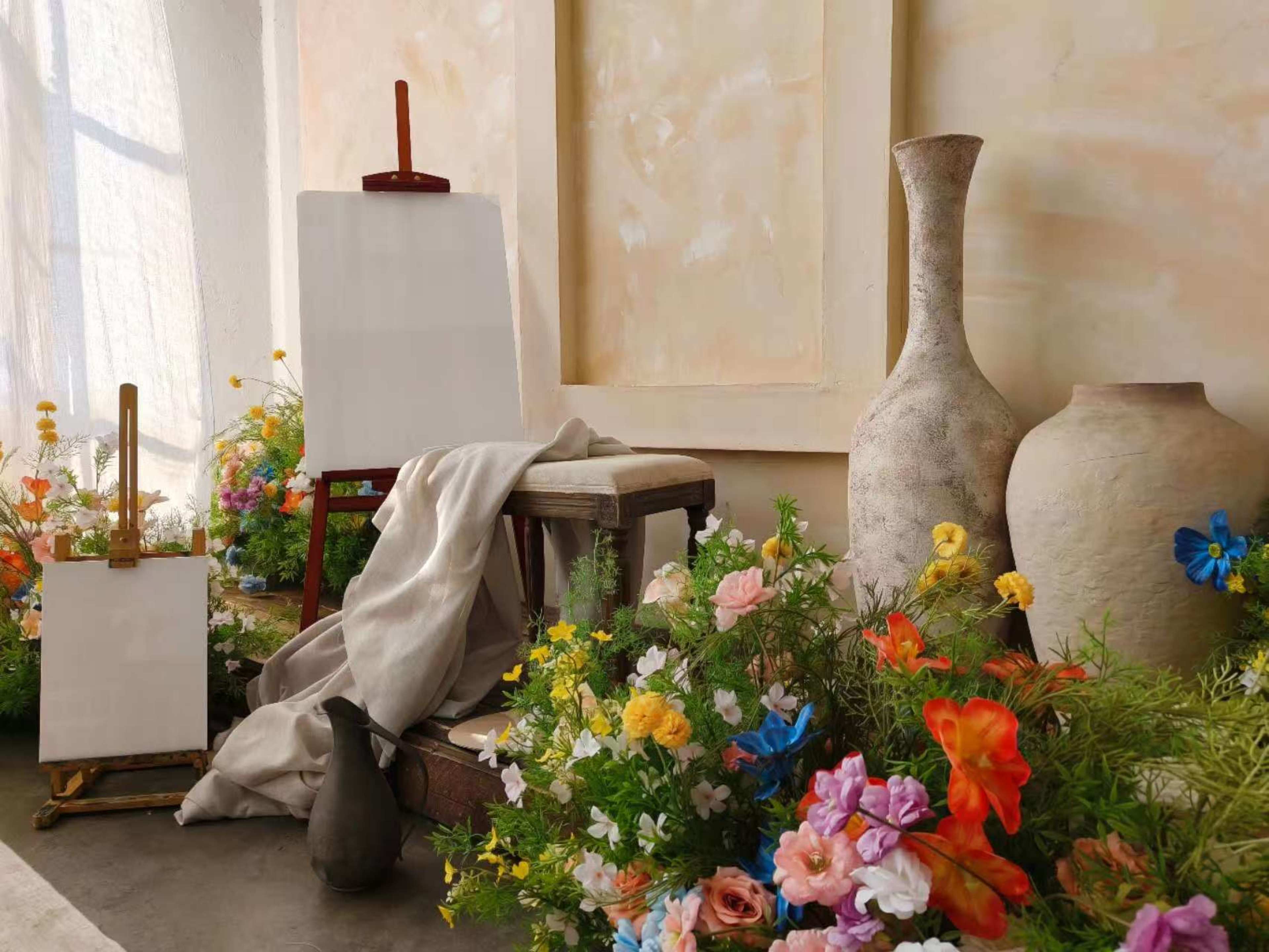 The image shows an artist's setup featuring blank canvases, a stool, a cloth draped over a chair, and vases surrounded by various colorful flowers.