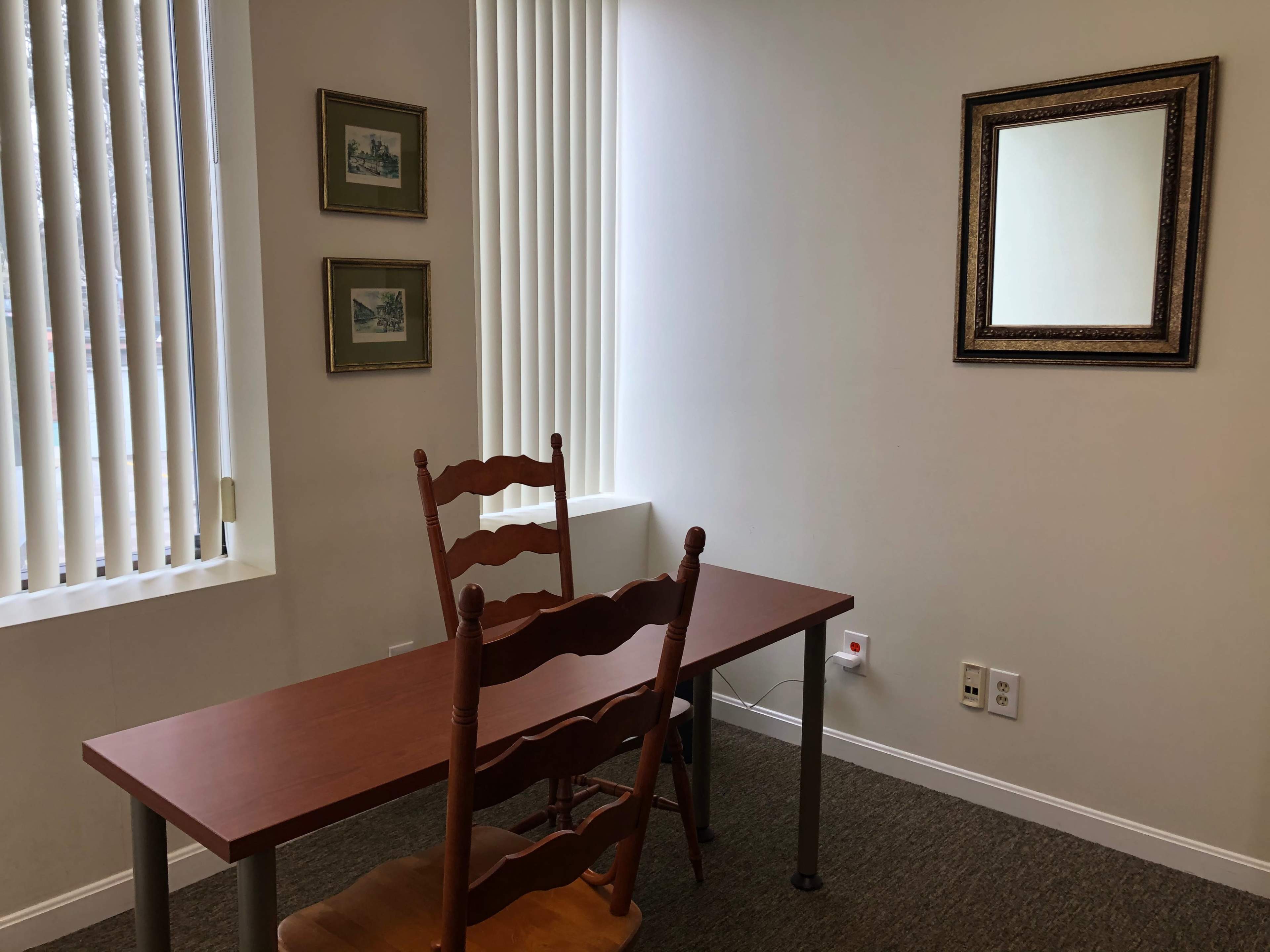 A small, minimally furnished room with a wooden table and two upright chairs, a mirror on the wall, and framed pictures.
