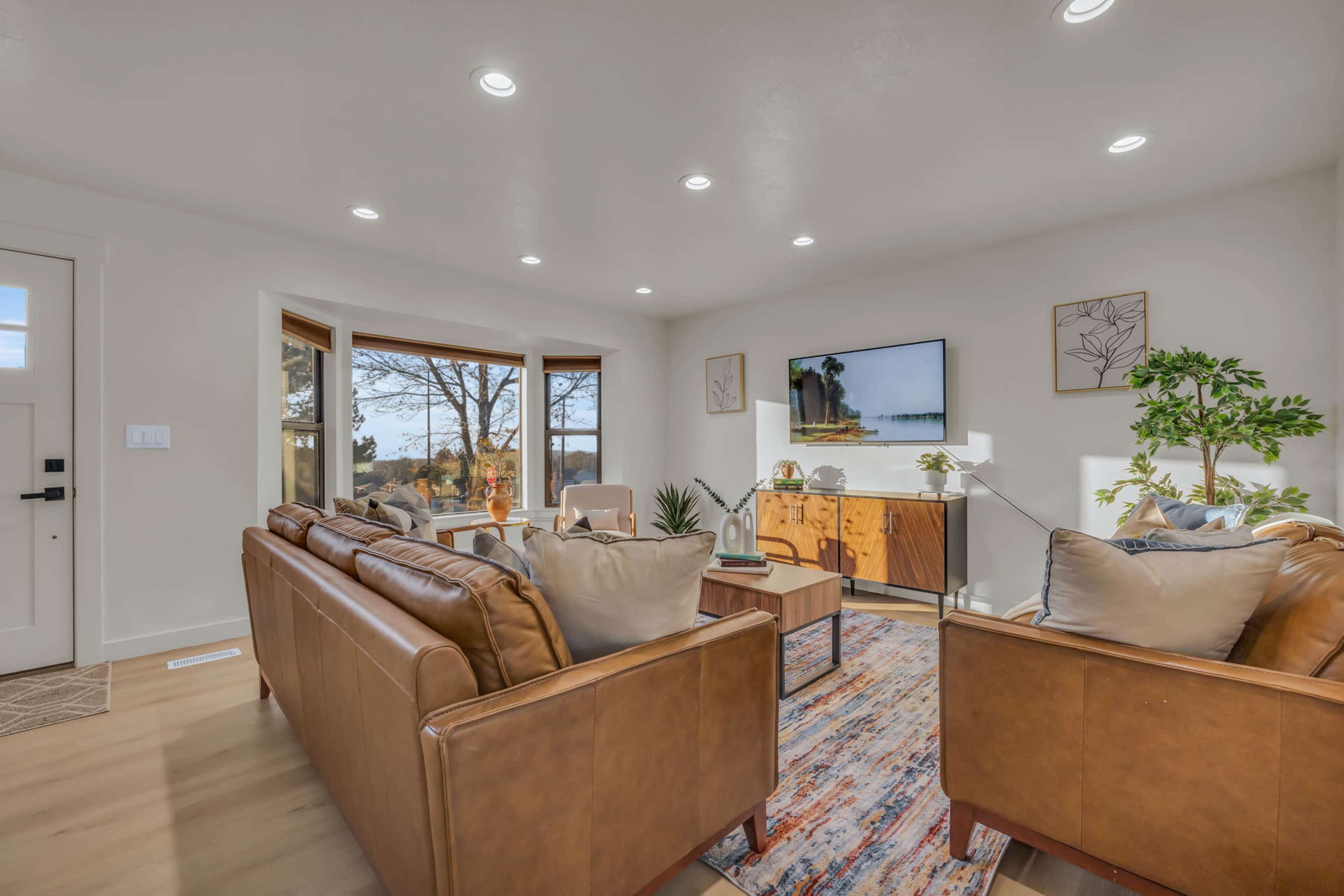 A modern living room features two brown leather sofas facing a wooden coffee table, with a television mounted on the wall and large windows providing natural light.