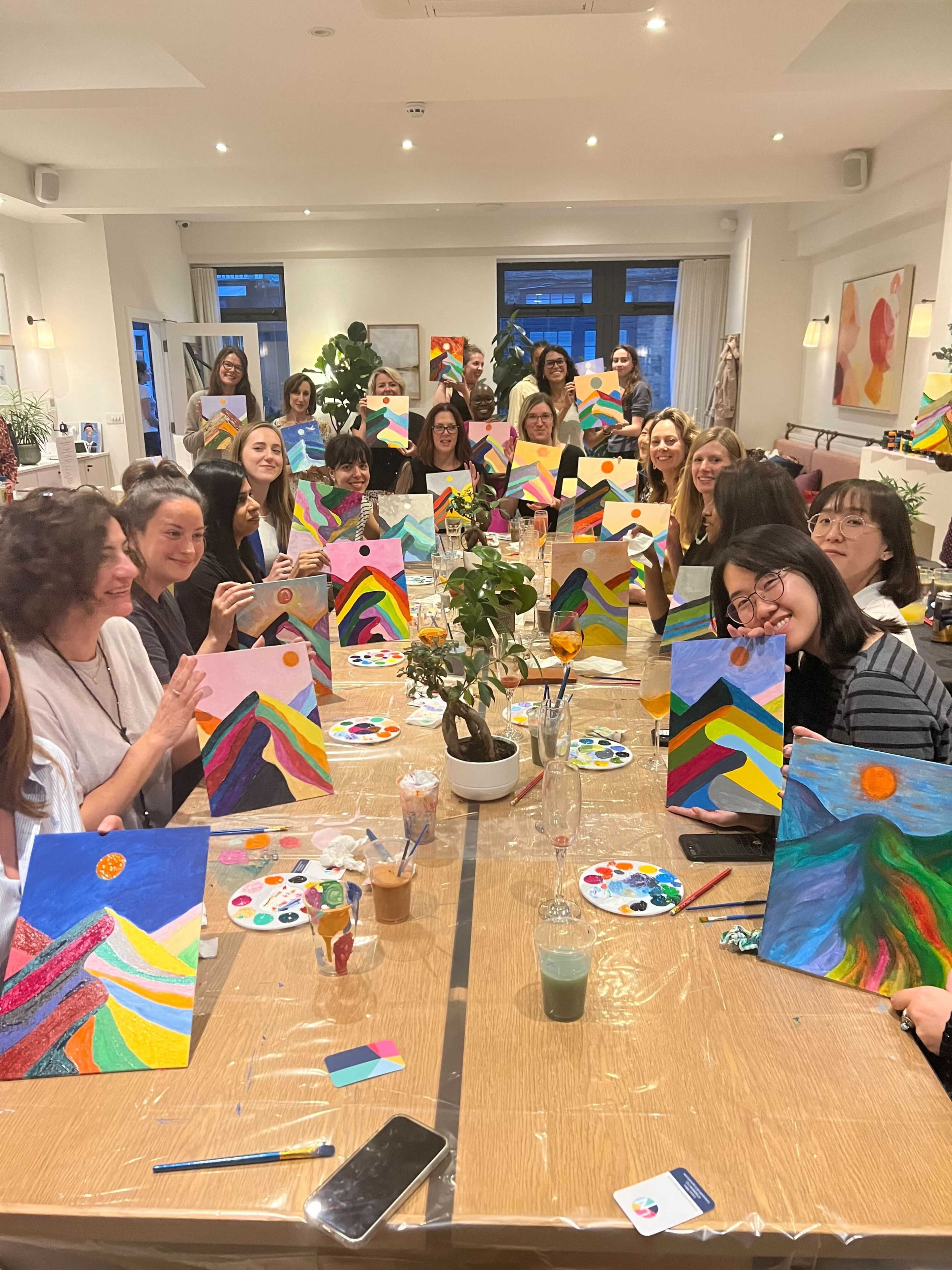 A group of people is sitting at tables in a bright room, each holding a painted canvas while surrounded by art supplies.