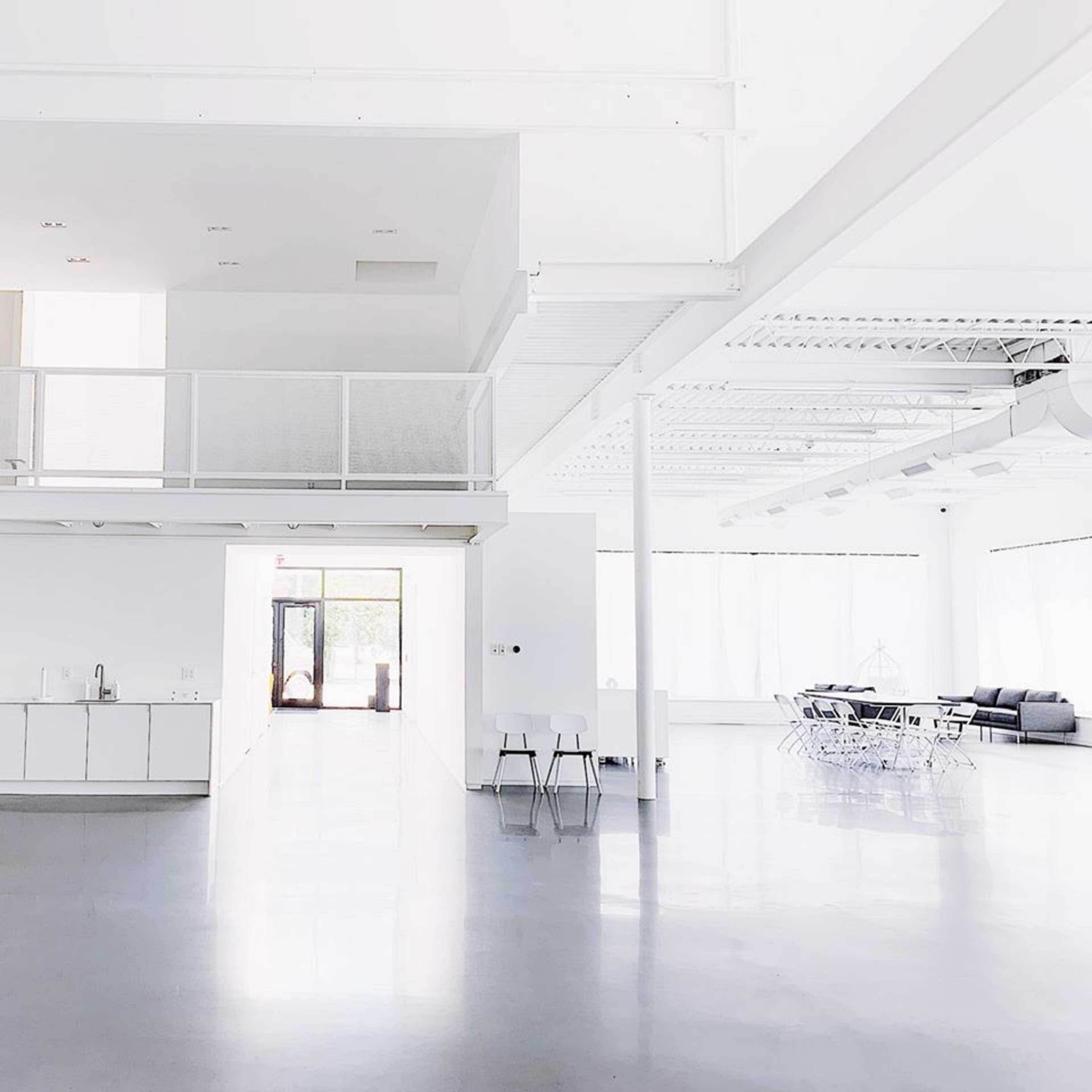 A spacious, modern interior with white walls, a gray floor, and minimal furnishings, including a kitchen area and a dining space.