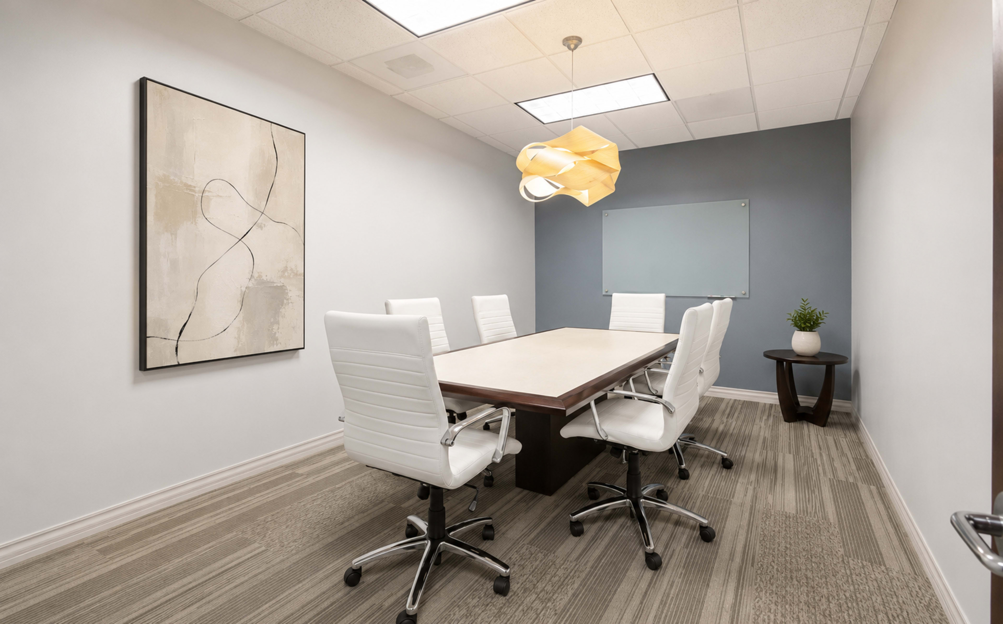 The image shows a modern conference room featuring a rectangular table surrounded by six white swivel chairs, with a light fixture above and a plant in a decorative pot.