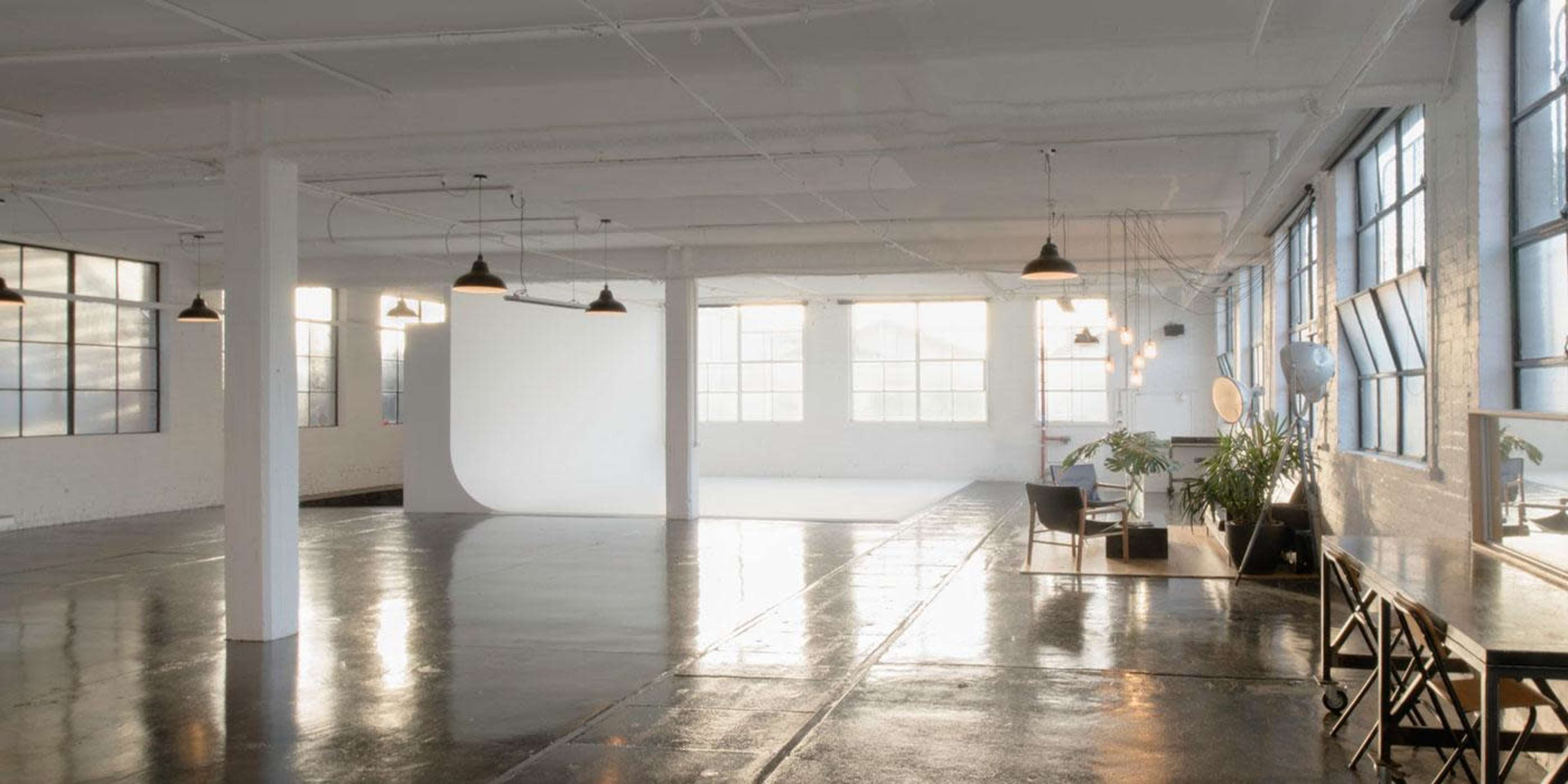 A spacious, well-lit studio with large windows, polished concrete floors, and minimal furnishings, including a table and chairs.