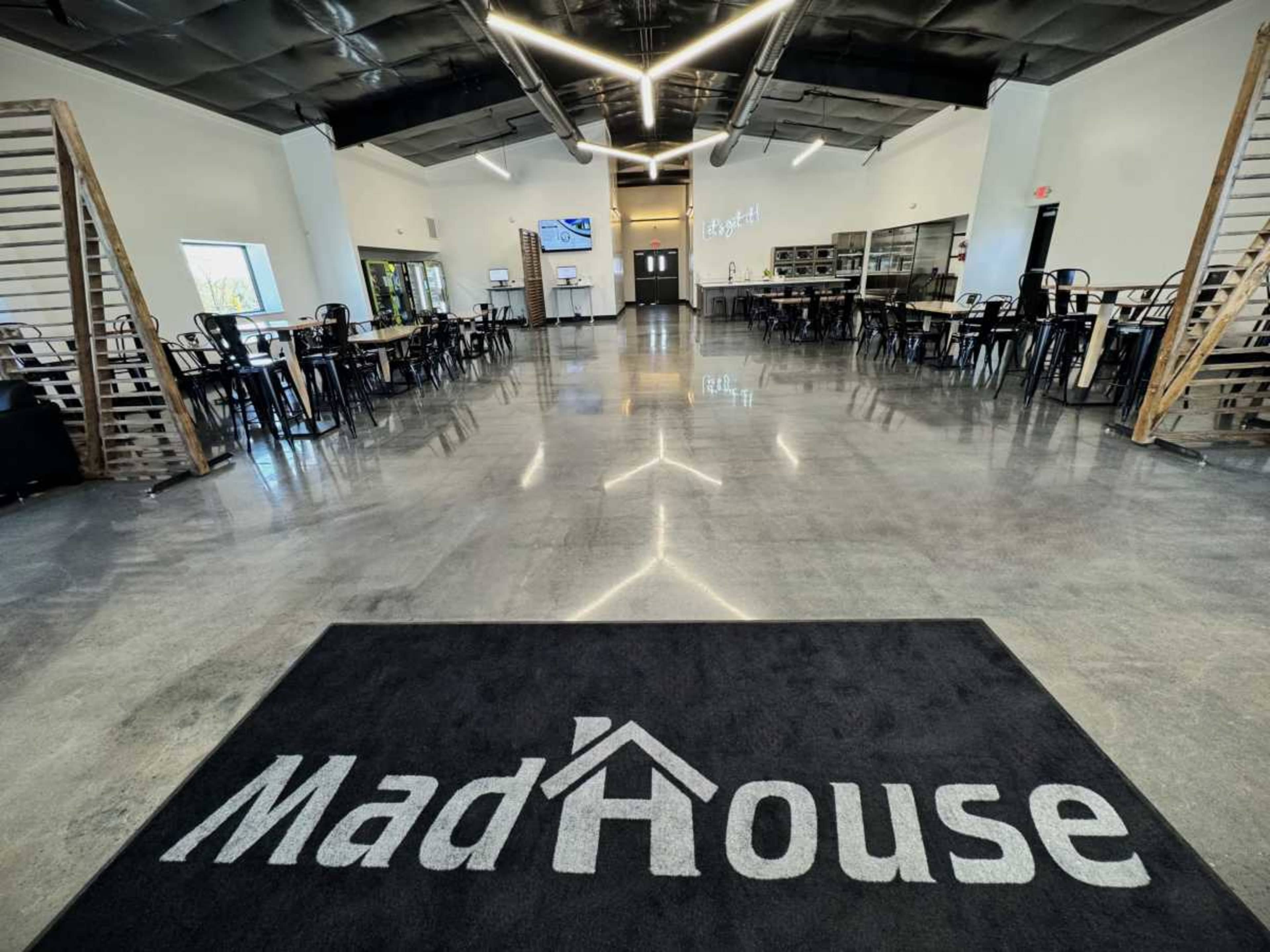 The interior of a modern dining space features a polished concrete floor, rows of black chairs and tables, and a prominent entrance rug with the name "MadHouse."