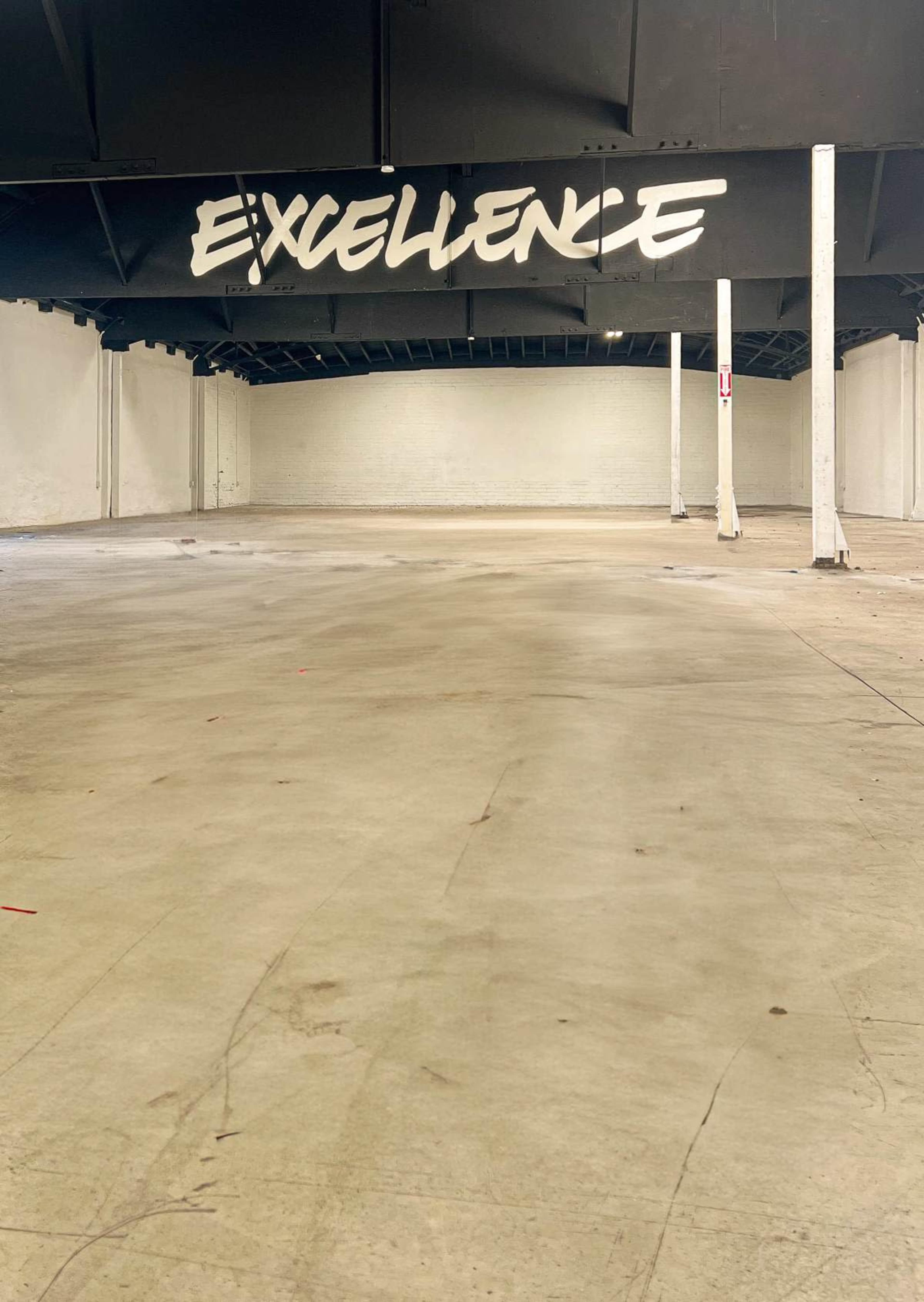 The image shows an empty warehouse space with white walls and a ceiling featuring the word "EXCELLENCE" painted in bold letters.