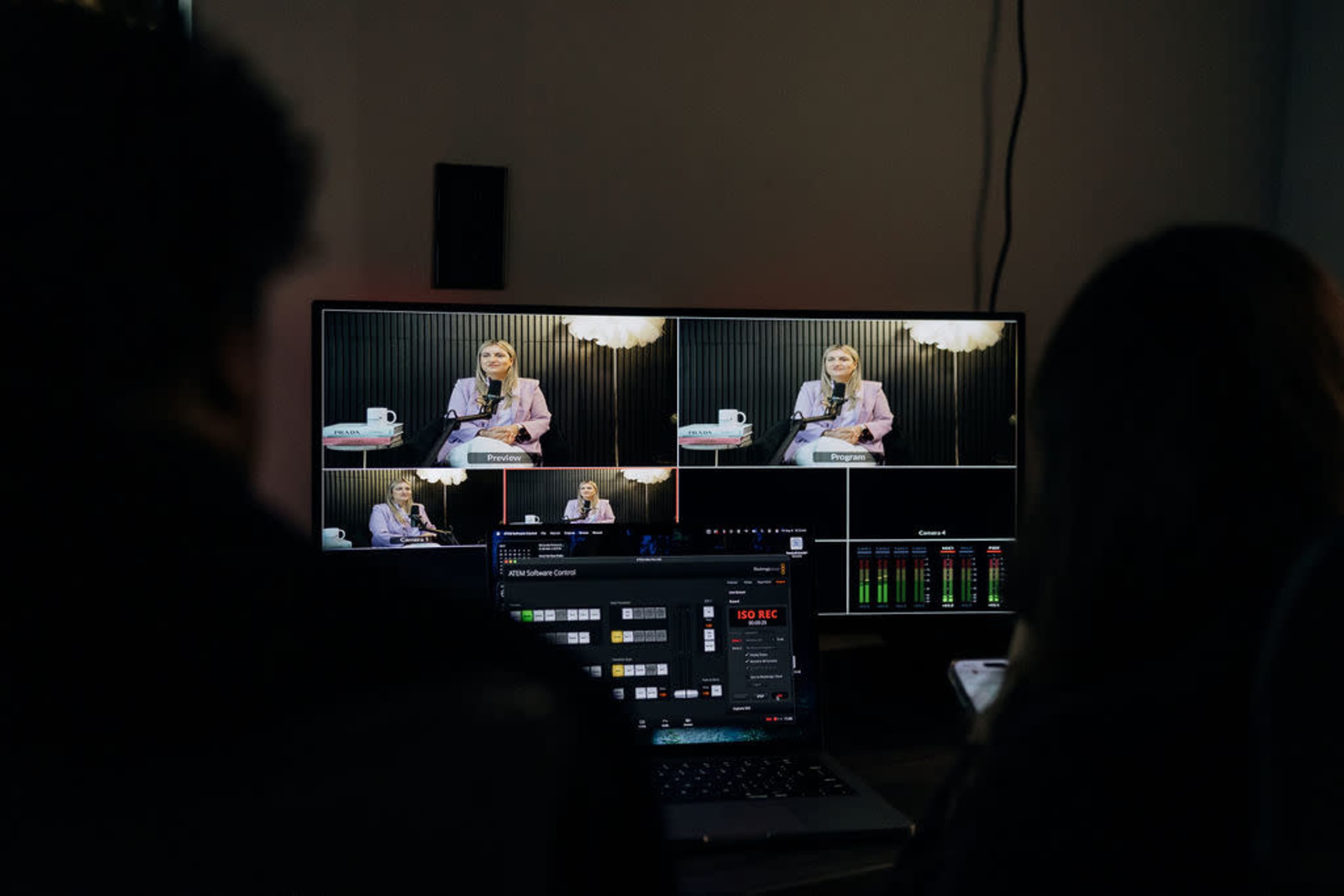 The image shows two people sitting in front of a multi-screen setup displaying a woman speaking into a microphone during a video production.