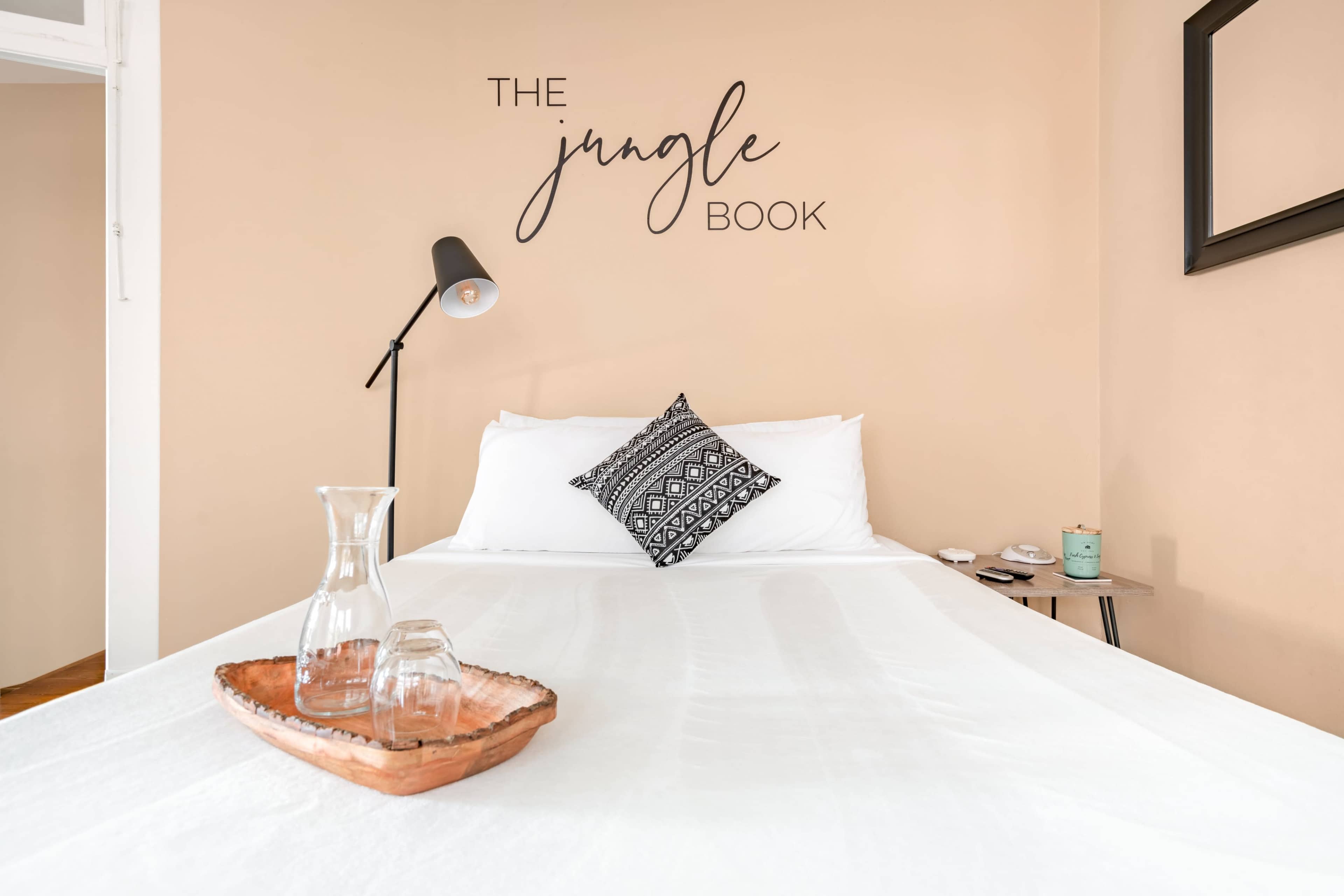 A neatly made bed with a decorative pillow, a glass water pitcher, and a wooden tray on a bedside table, against a beige wall featuring the words "THE JUNGLE BOOK."