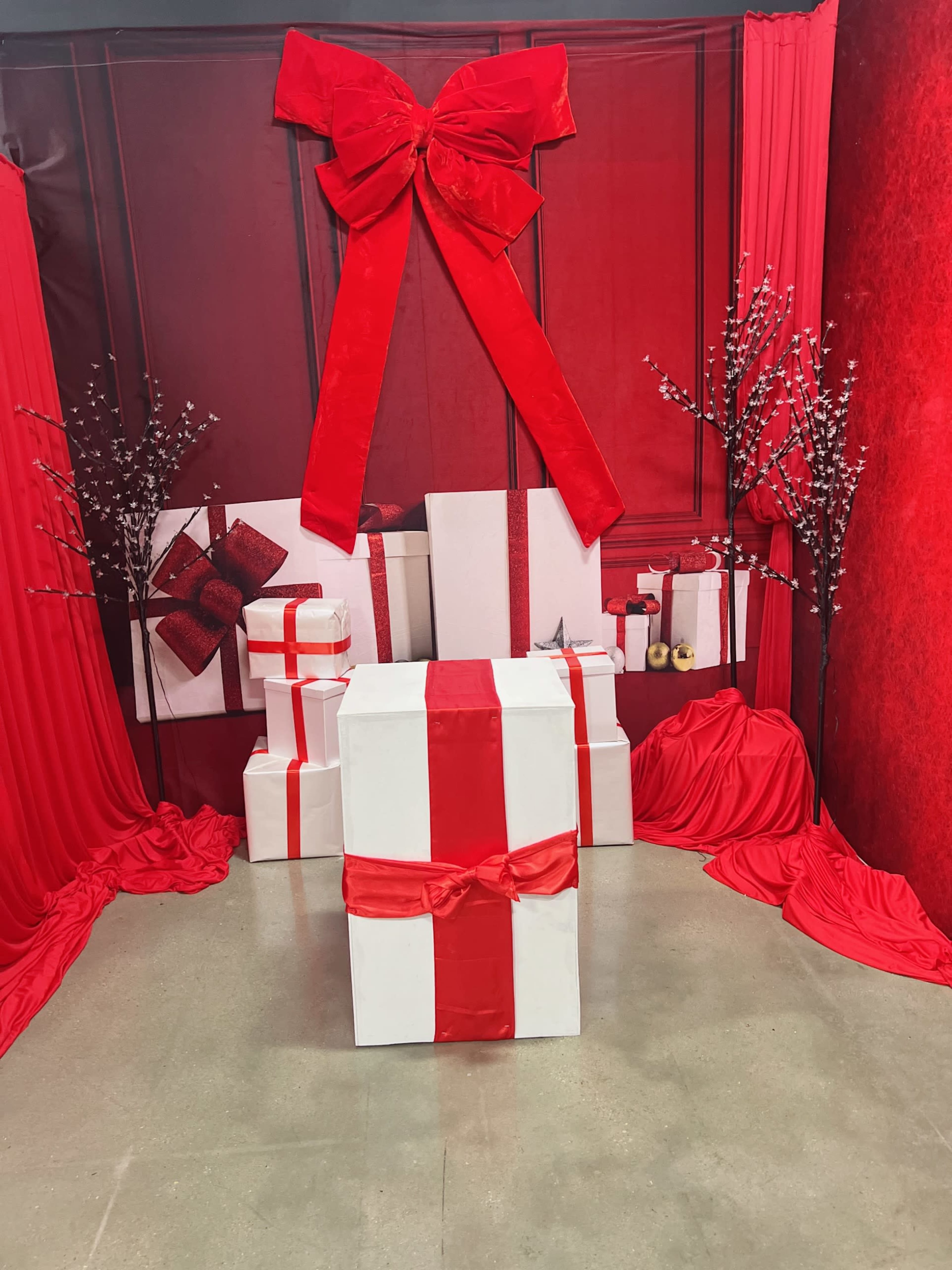 The scene features a red and white themed display with large gift boxes, a big bow, and decorative branches set against a matching backdrop.