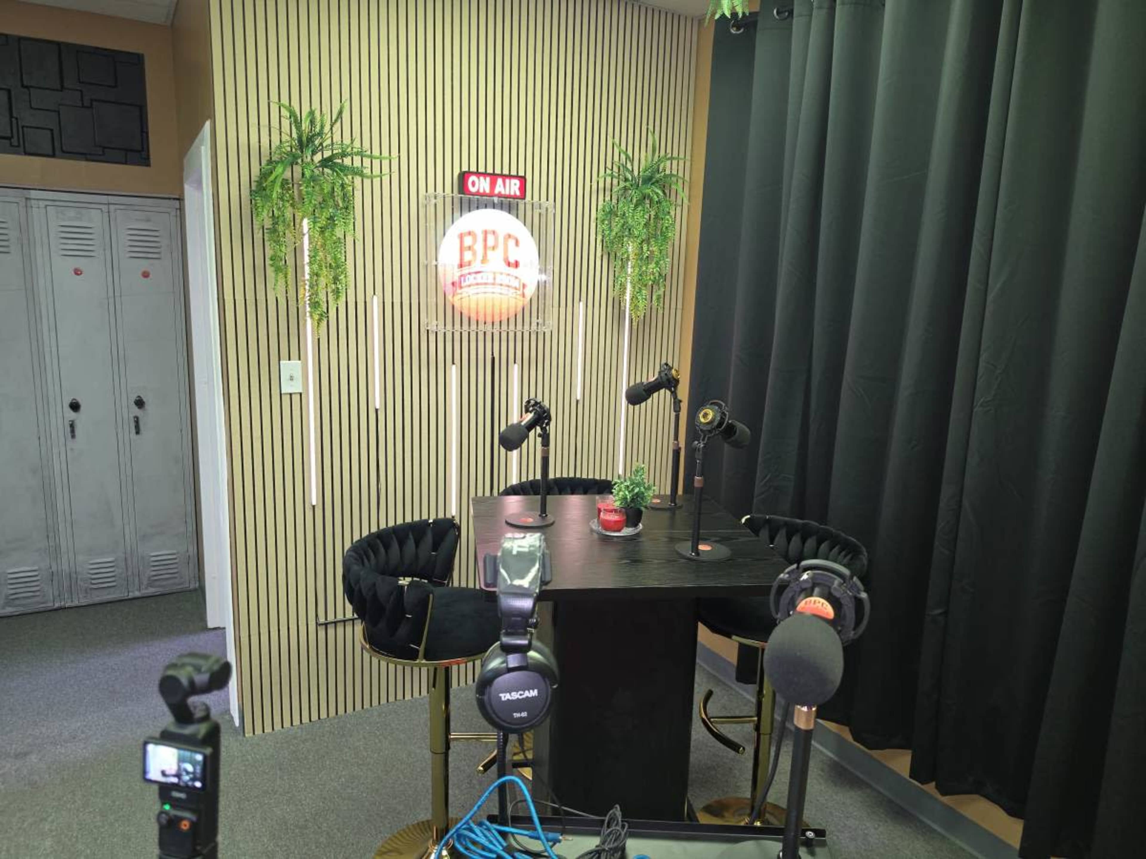 The image shows a small recording studio setup with a table, two microphones, a potted plant, and an "ON AIR" sign on the wall.