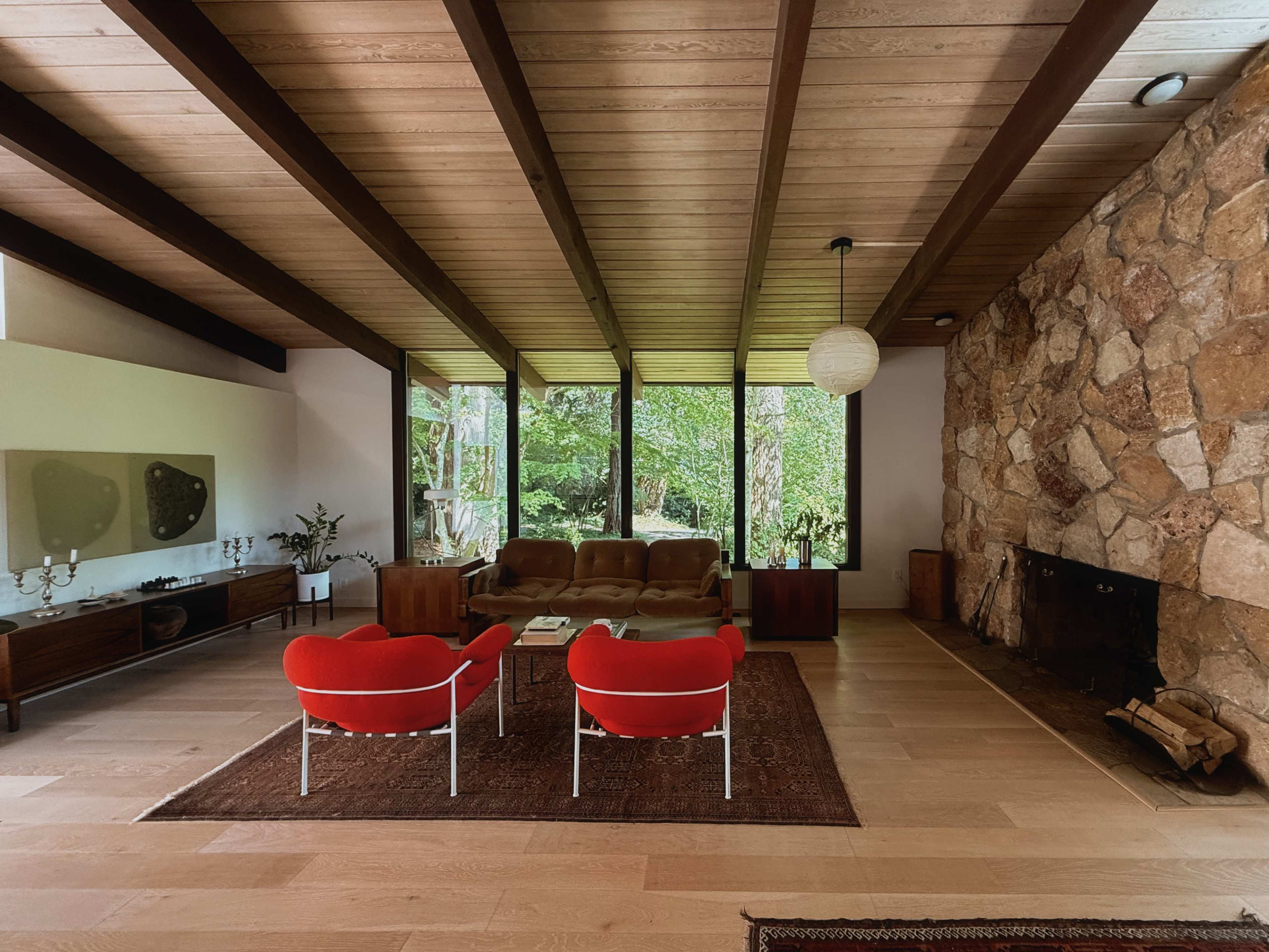 The image shows a modern living room with wooden beams, a stone fireplace, large windows overlooking greenery, and red chairs positioned around a coffee table on a rug.