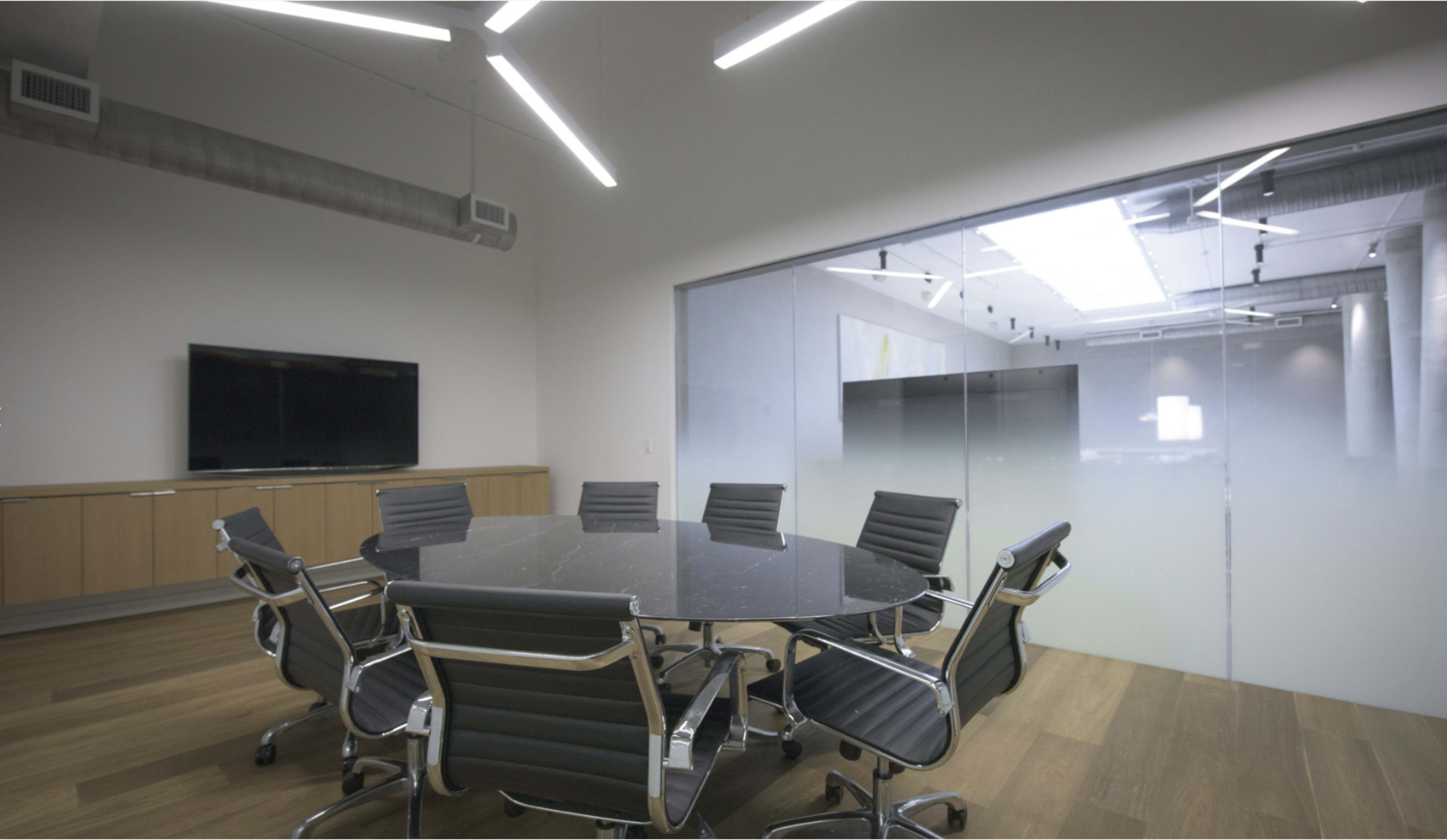 The image depicts a modern conference room featuring a round black table surrounded by ergonomic chairs, with a large screen on the wall and a glass wall separating it from an adjoining space.
