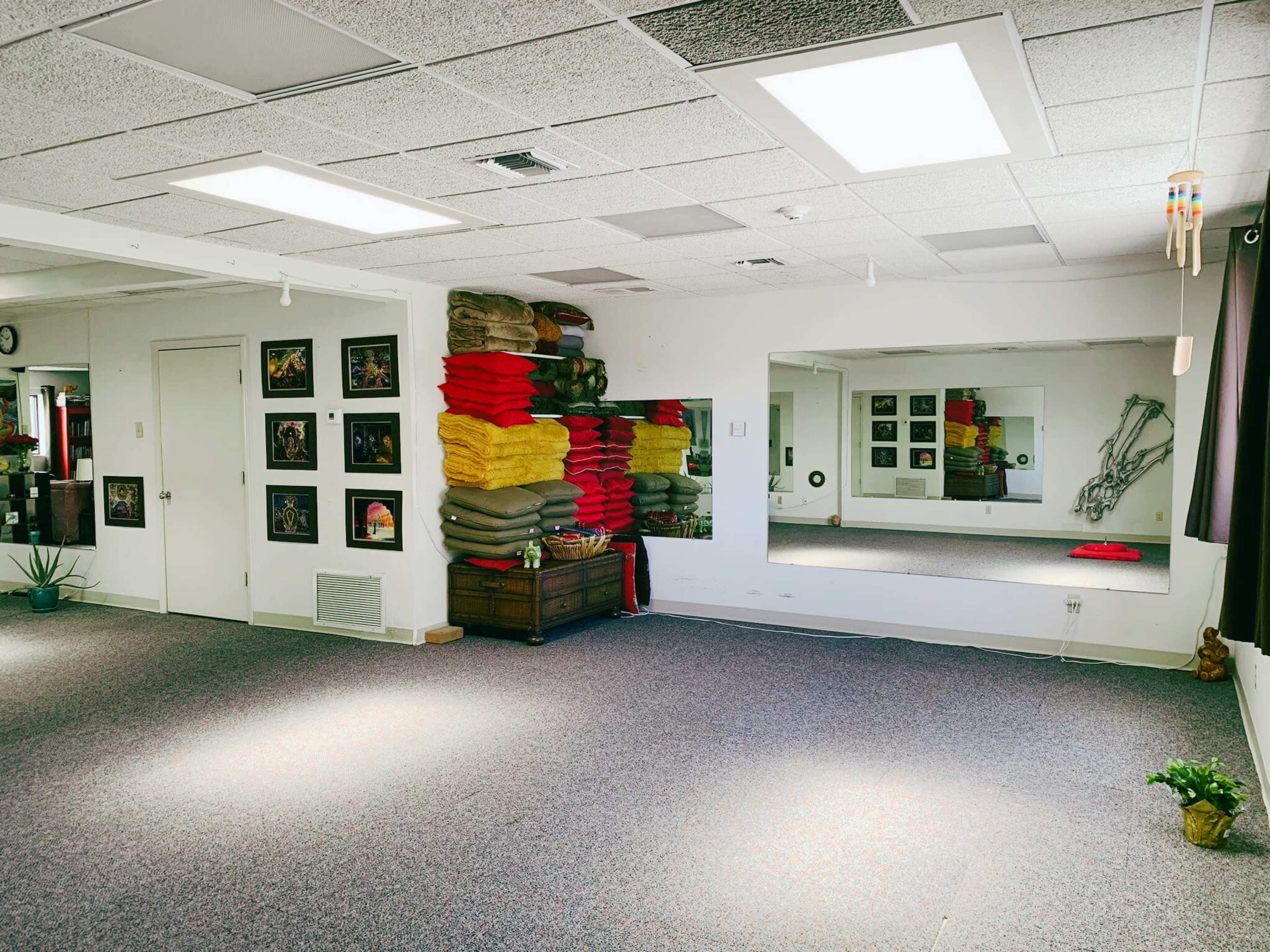 A spacious room with carpeted flooring, featuring colorful stacks of blankets against one wall, a large mirror, and framed artwork hanging on another wall.