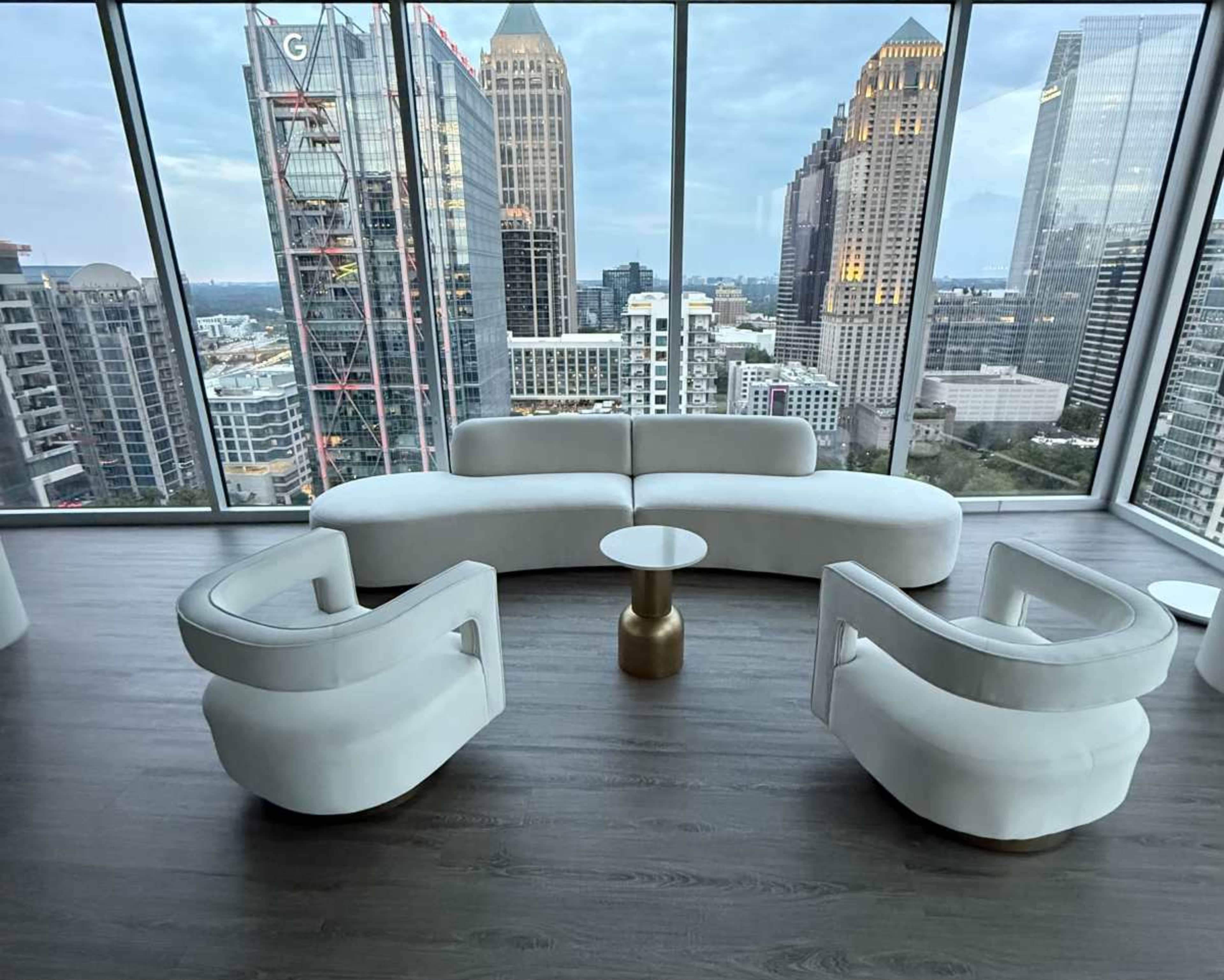 The image shows a modern seating area with a curved white sofa and two armchairs surrounding a small, round table, set against a panoramic city skyline.