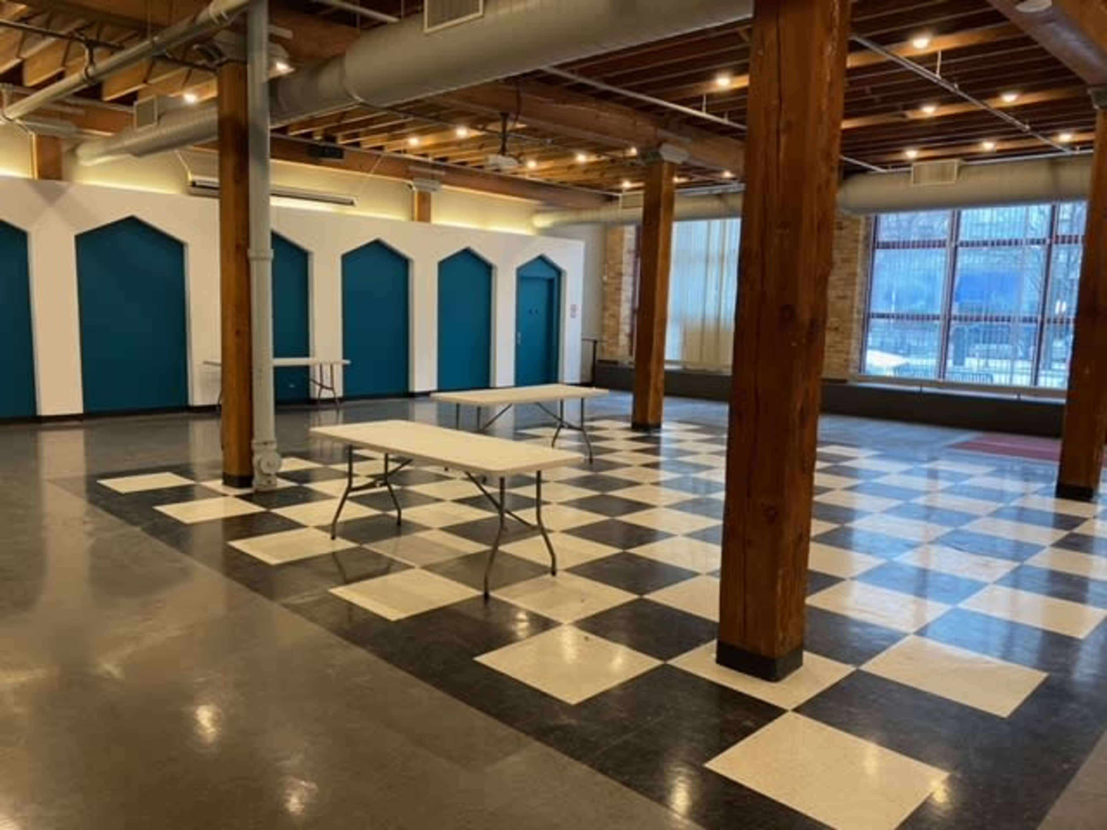 A large, open interior space with checkered flooring, wooden pillars, and several white folding tables arranged in front of teal doors.