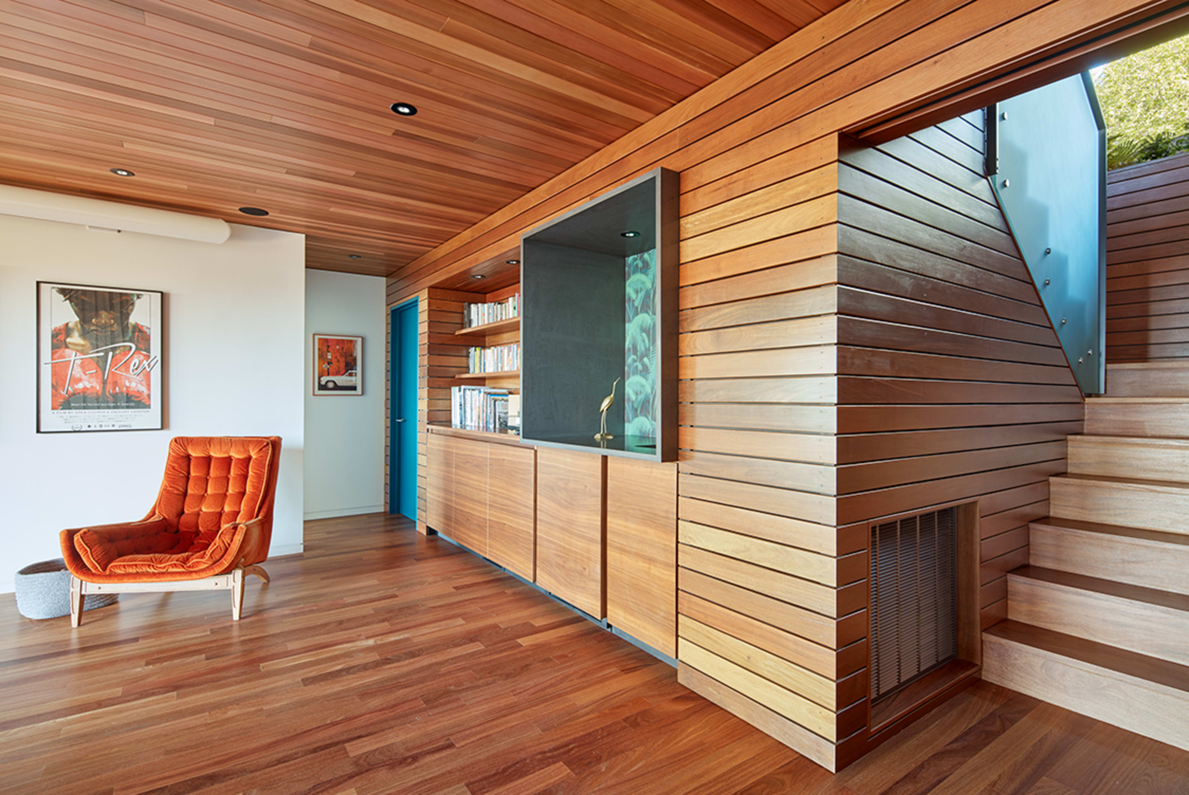 A modern interior features wooden walls, a staircase, and a cozy orange chair beside a bookshelf, with a framed poster on the wall.