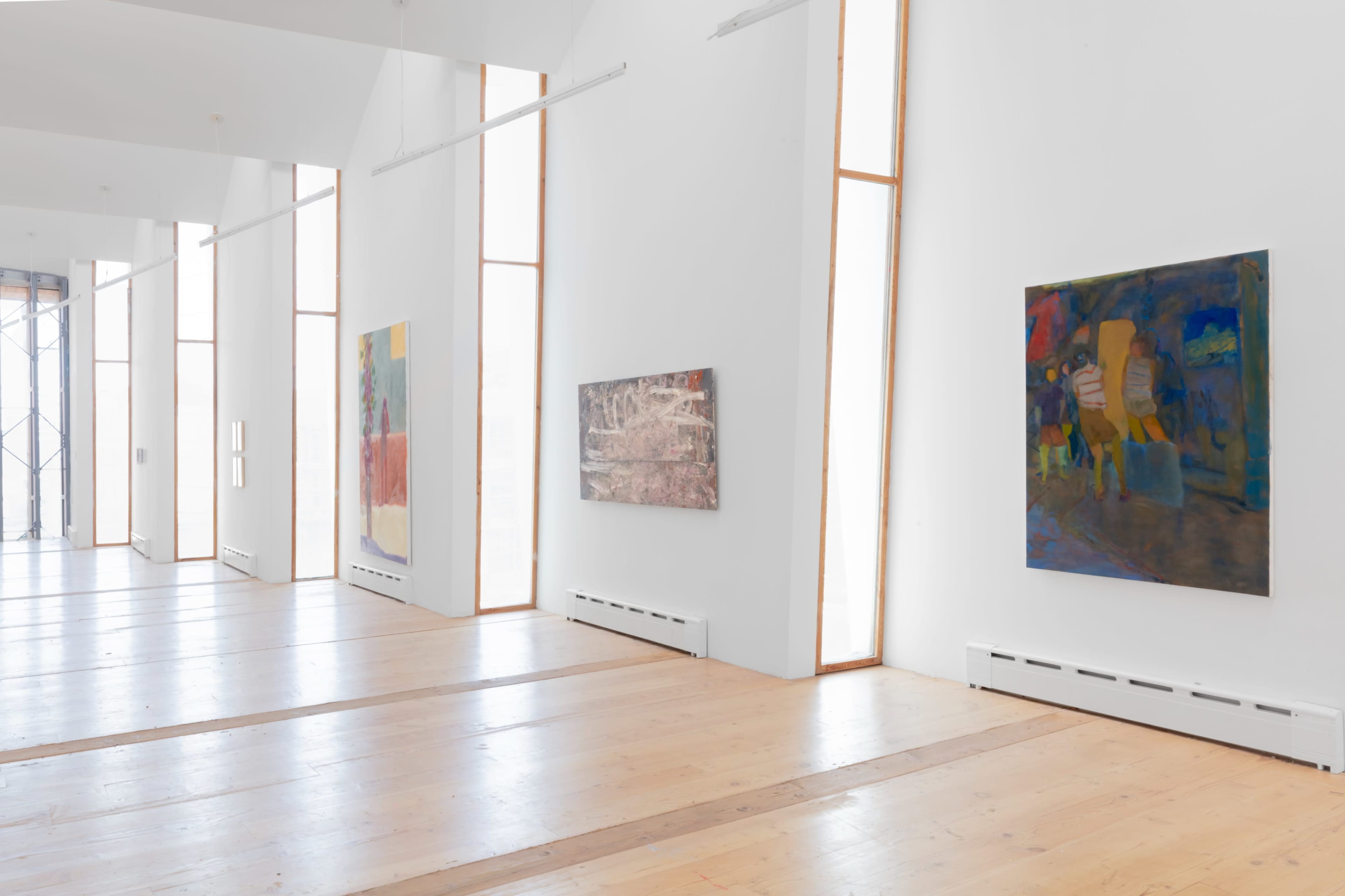 The image shows a bright gallery space with large windows and several colorful paintings hanging on the white walls.