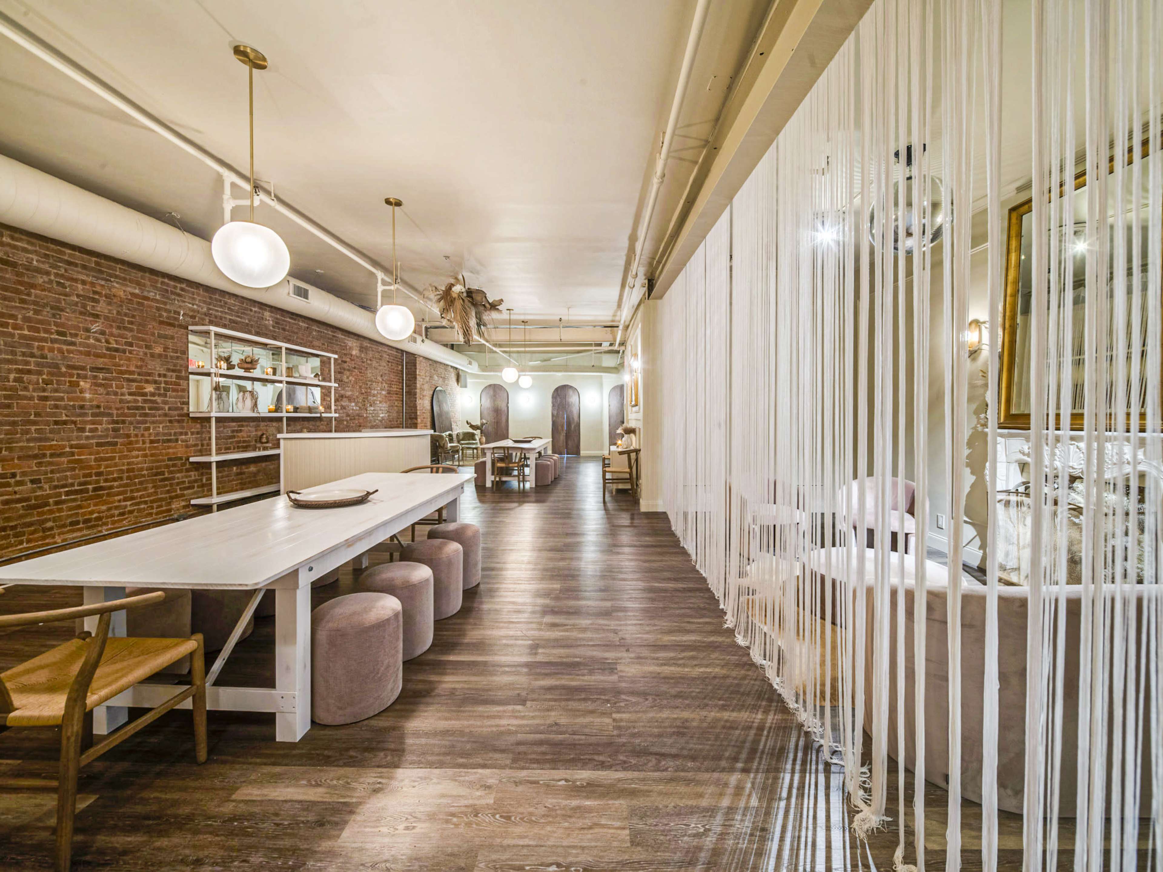 A modern restaurant interior featuring a long white table, round stools, exposed brick walls, and dividers made of hanging threads.
