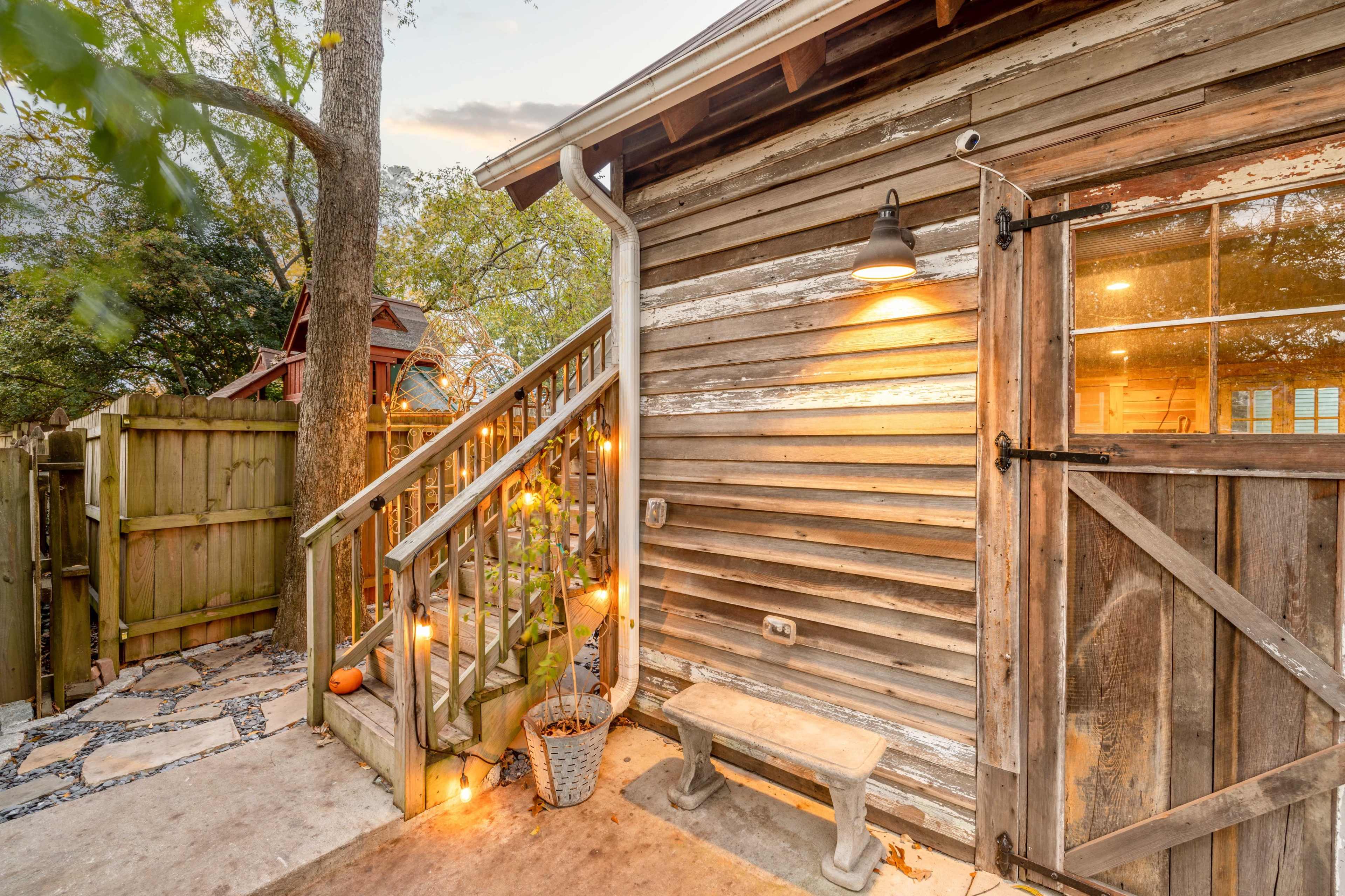 A rustic wooden exterior of a small building with a staircase leading up, illuminated by a wall-mounted light, and surrounded by greenery and garden decor.
