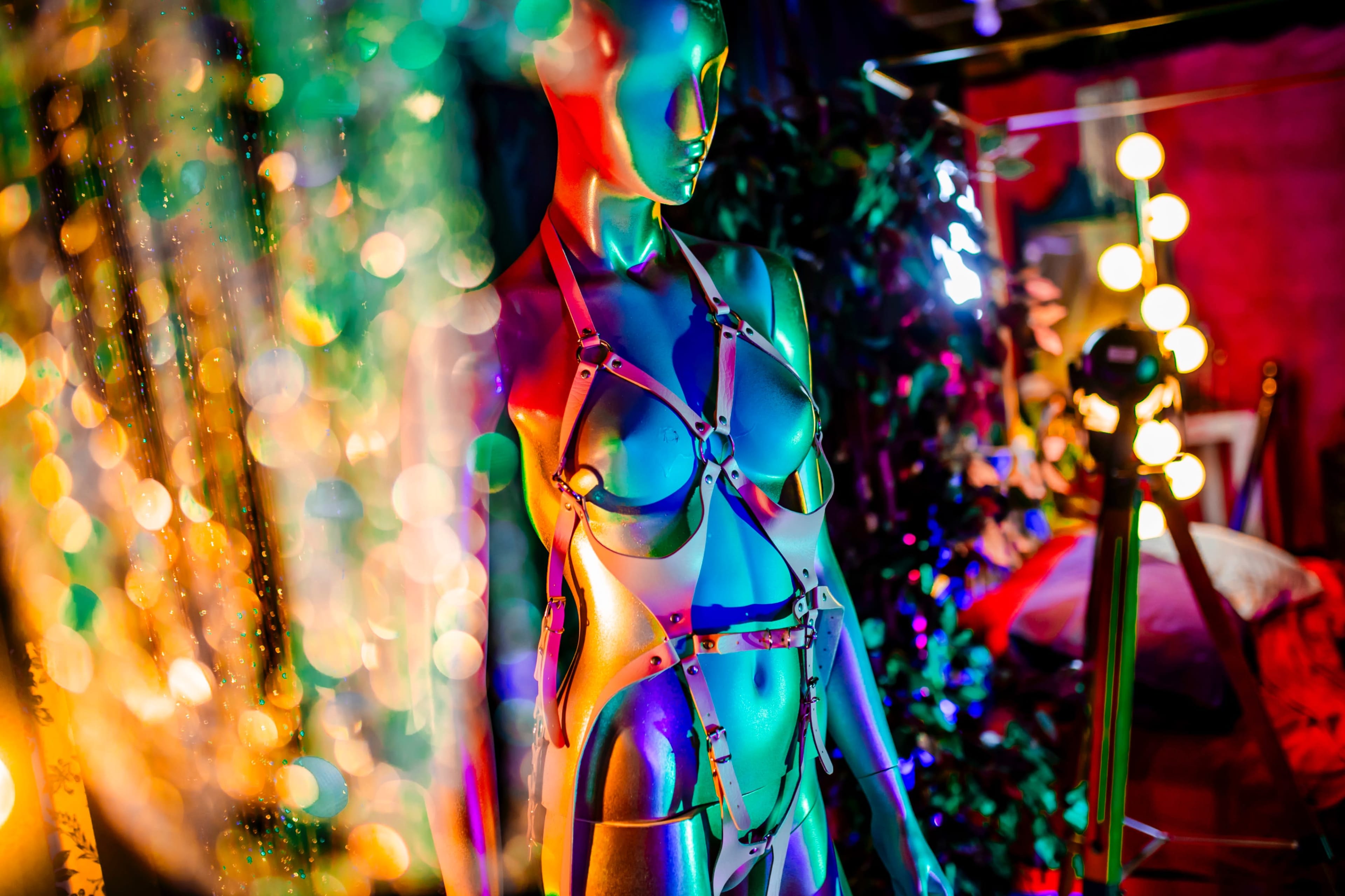 A metallic mannequin stands under colorful lights, adorned with a harness, amidst a backdrop of vibrant decorations.