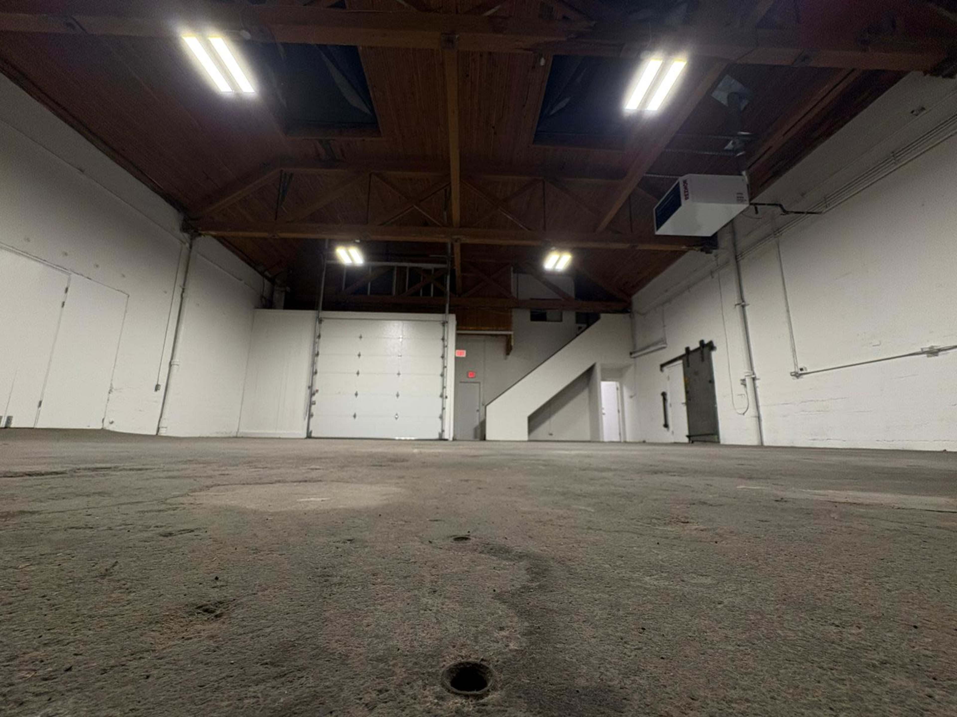 The image shows a spacious, empty interior of a warehouse with wooden beams, concrete flooring, and fluorescent lighting.