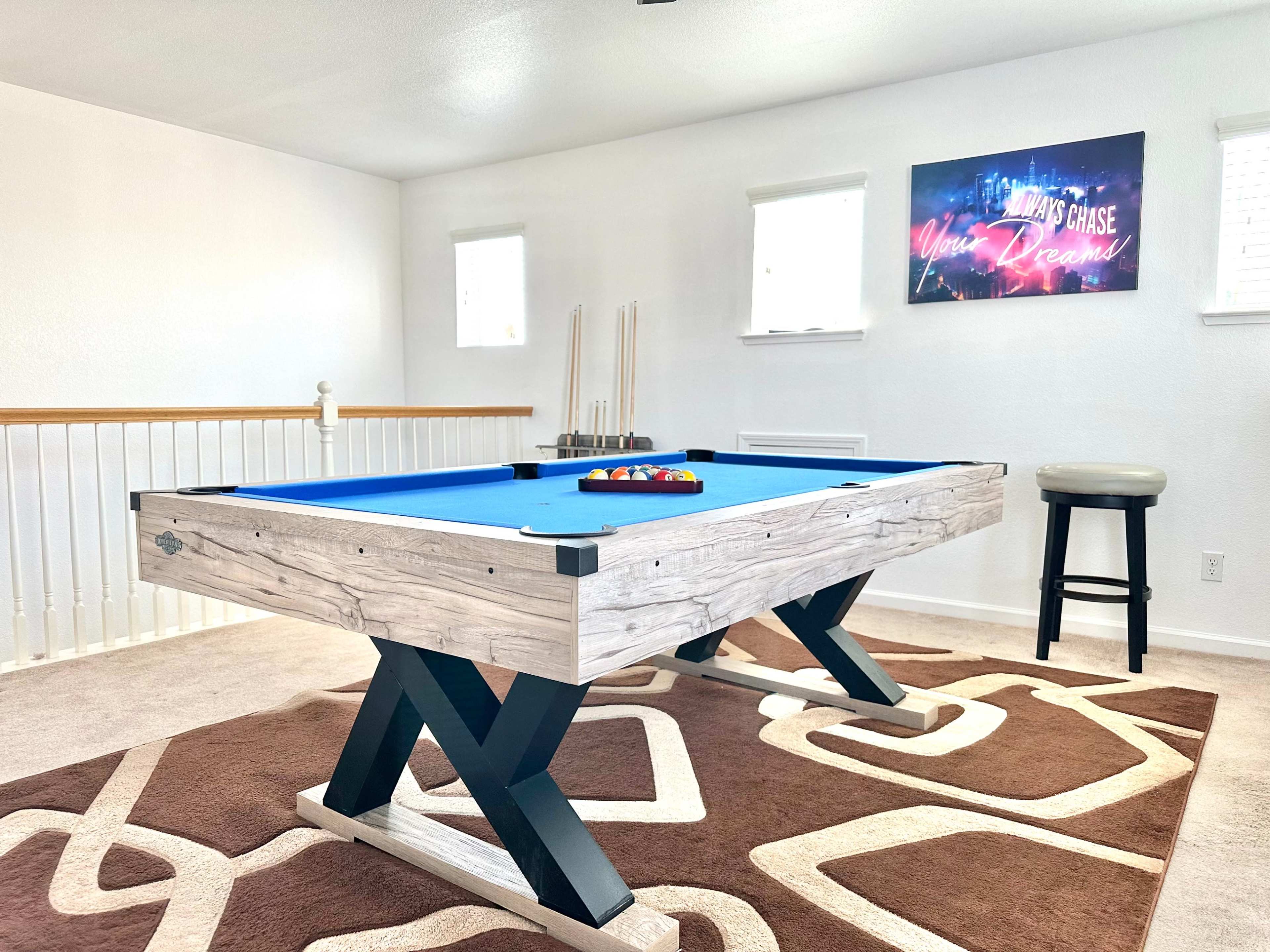 A spacious room with a blue pool table in the center, framed by white walls and two windows, and a brown patterned rug underneath.