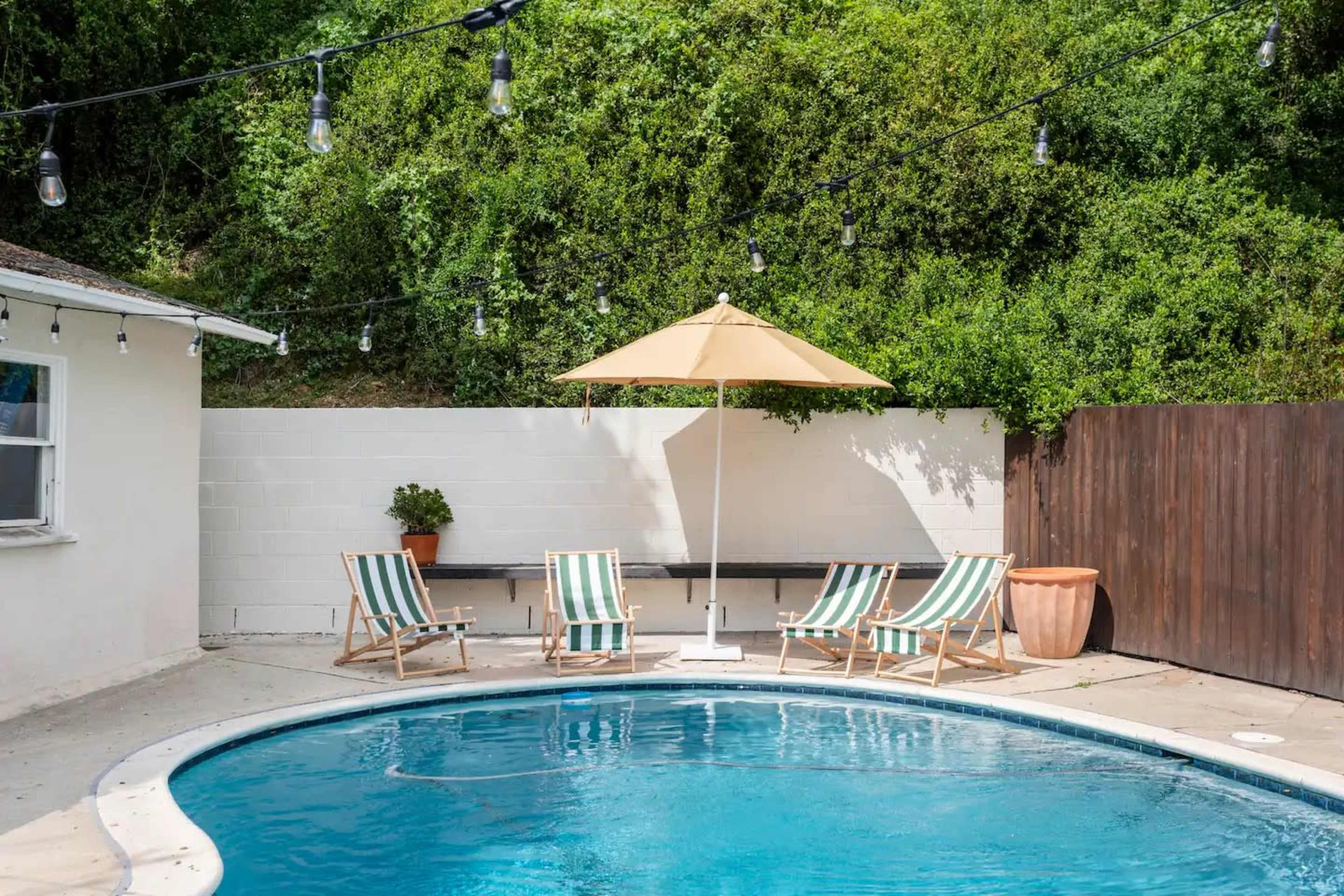 A swimming pool is surrounded by striped lounge chairs and an umbrella, with greenery and string lights in the background.
