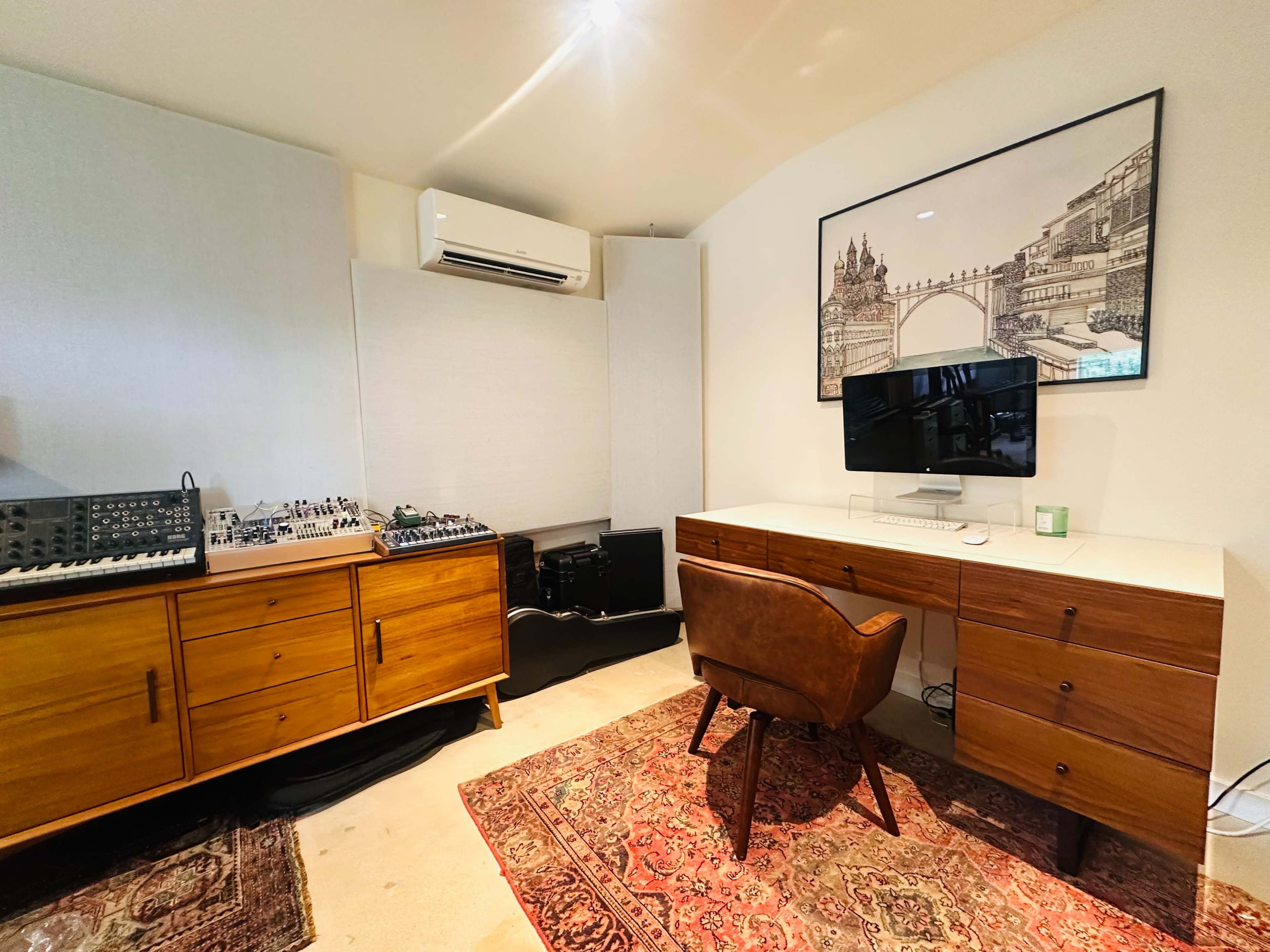 The image shows a music studio featuring a work desk with a computer and a chair, surrounded by audio equipment, storage cabinets, and a patterned rug.