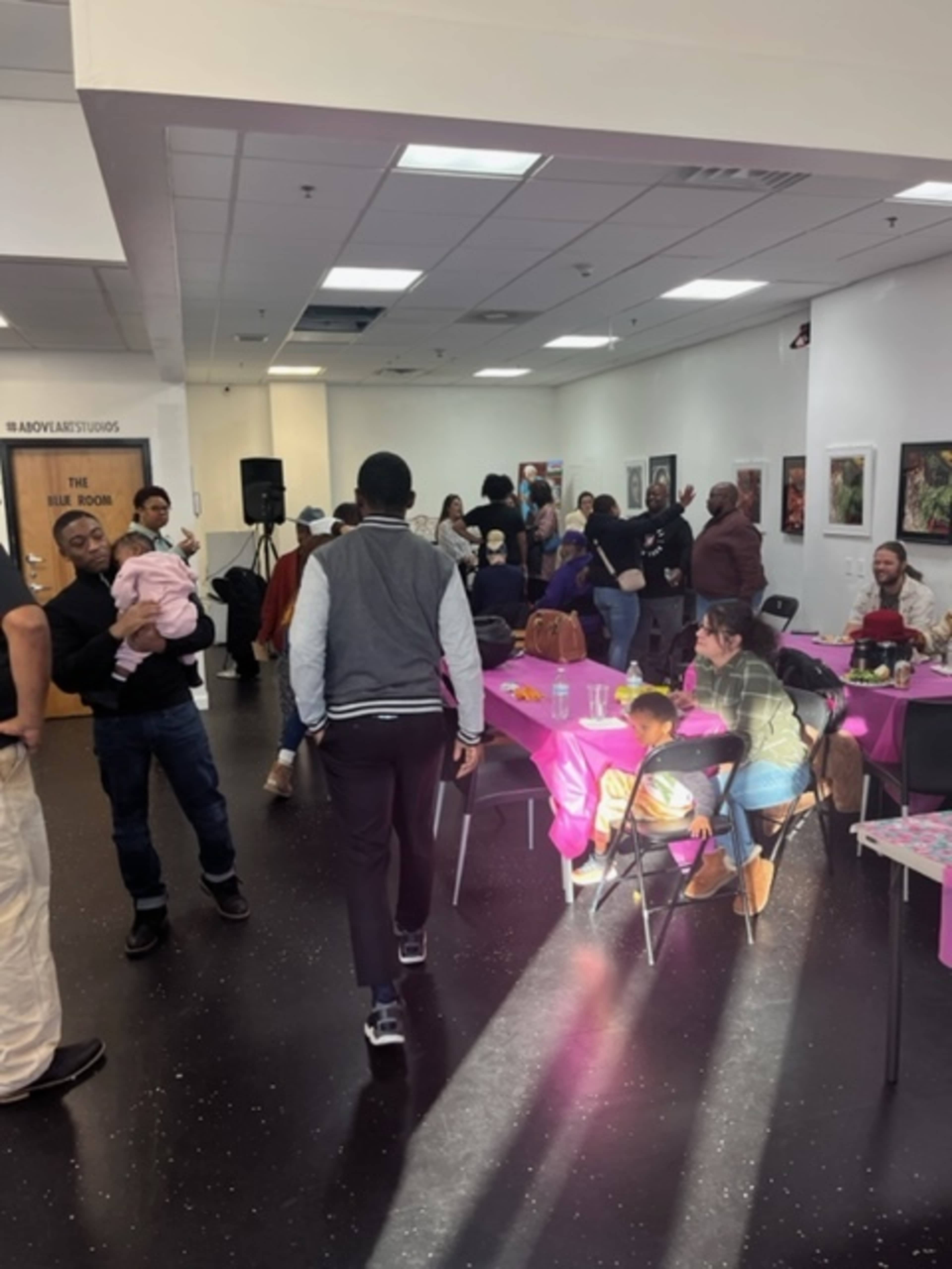 A diverse group of people gathers in a room with tables set for a social event, while some attendees mingle and others are seated.