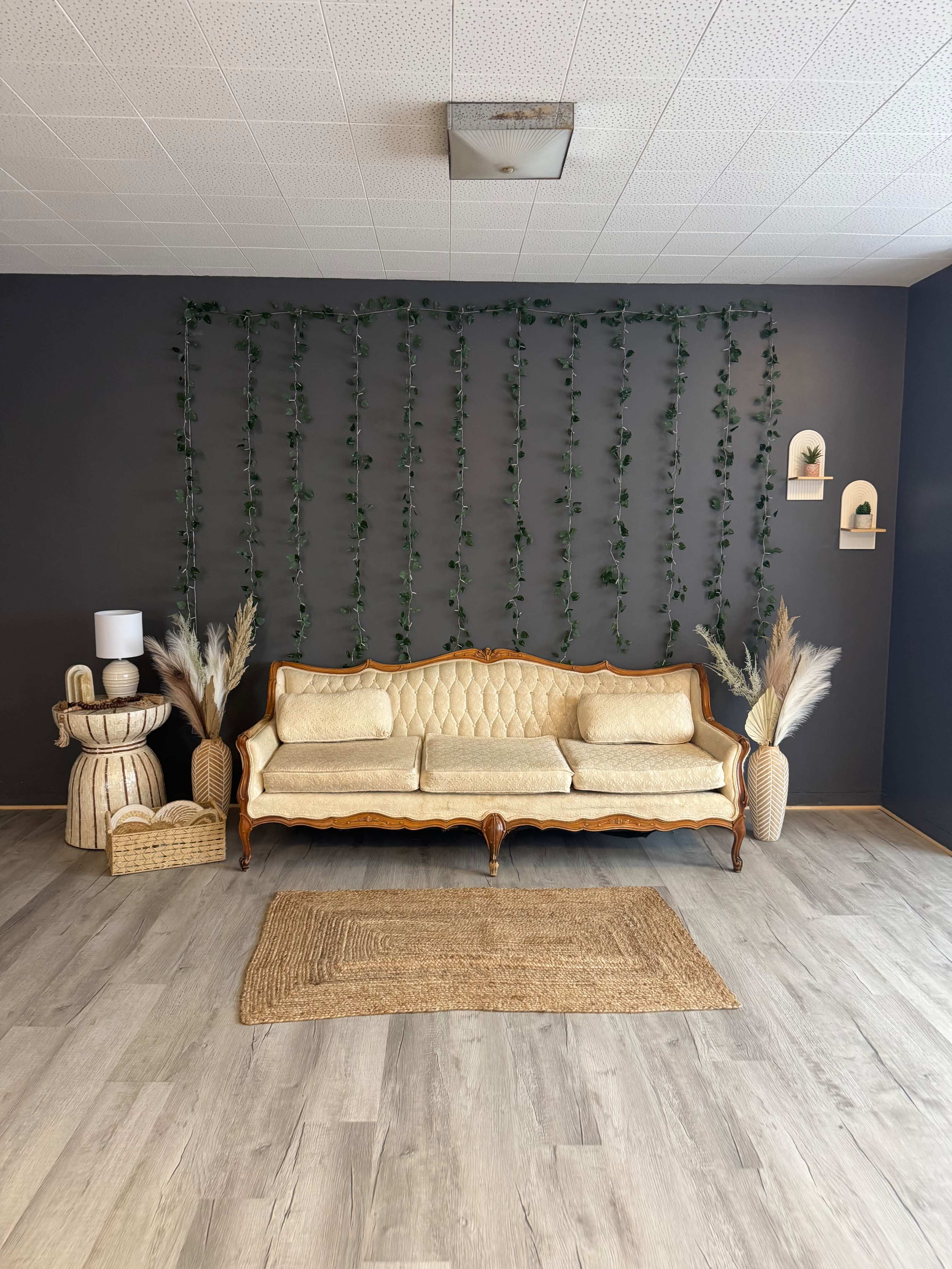 The image features a vintage-style, cream-colored sofa against a dark wall adorned with vertical green vines, flanked by decorative plants in baskets and wall-mounted shelves.