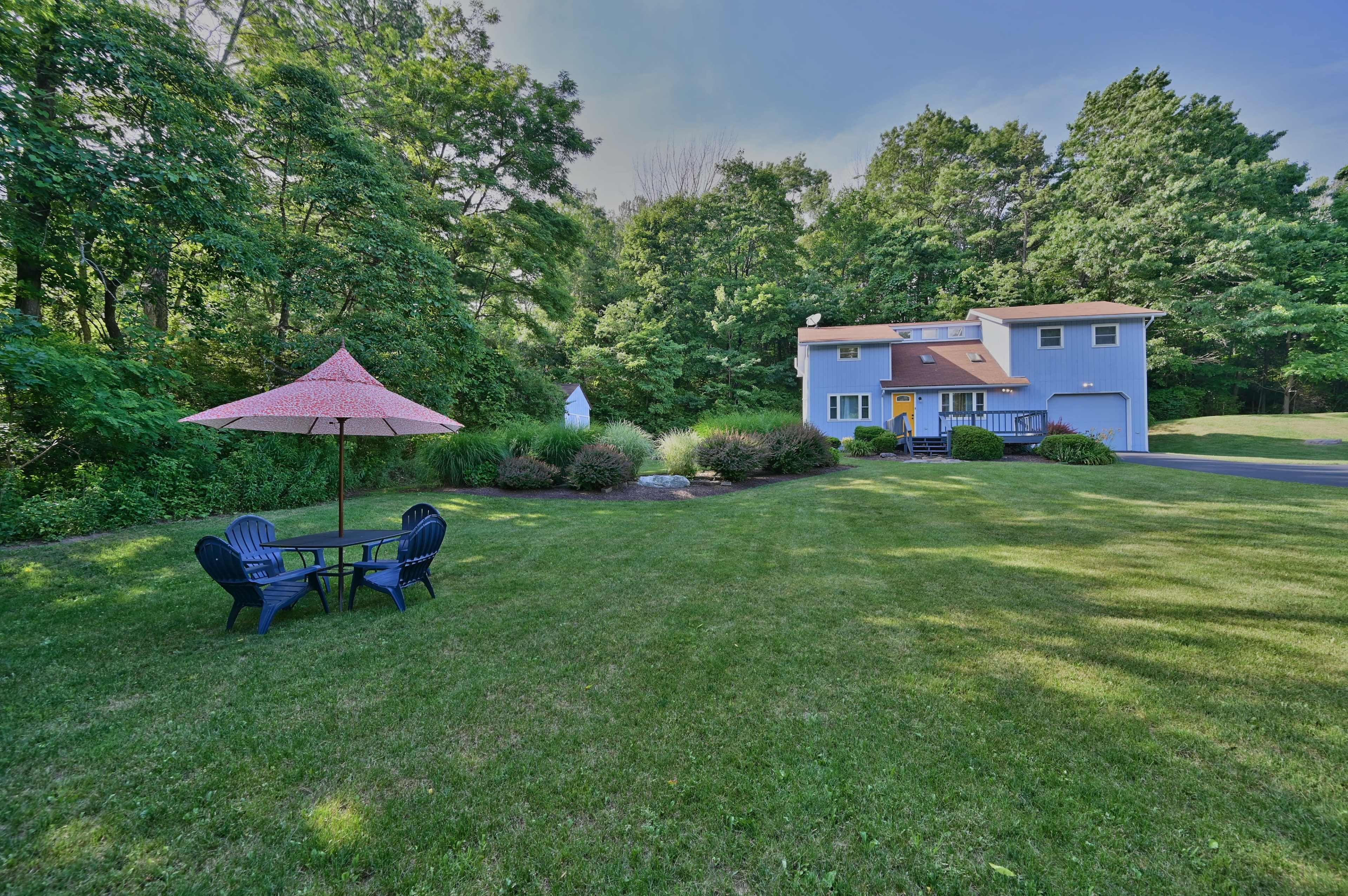 A blue two-story house is surrounded by trees, with a green lawn featuring a round table and chairs underneath a pink umbrella.