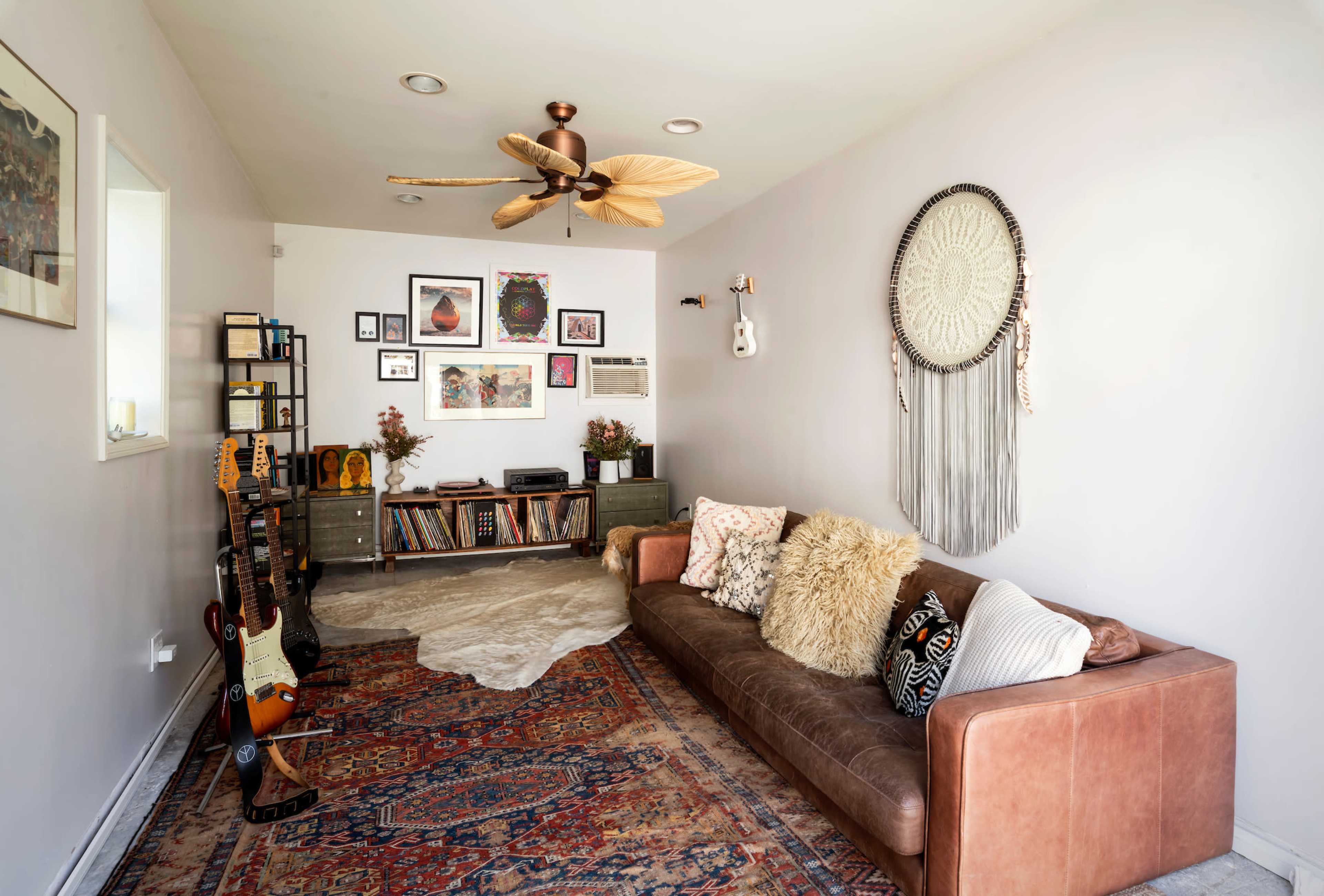 The room features a leather sofa, a patterned rug, and a mix of decor, including guitars, shelving, and wall art.