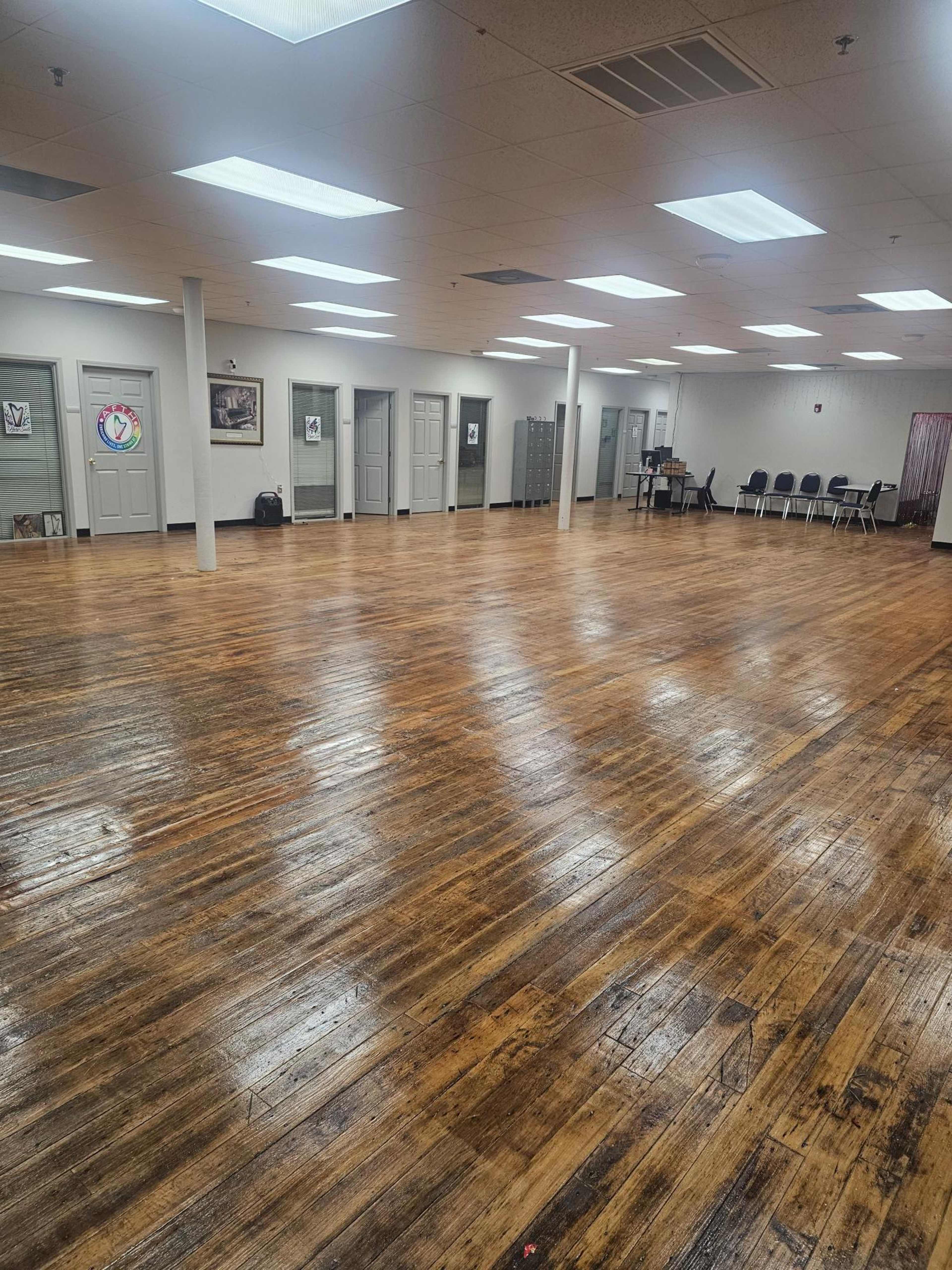 The image shows a spacious interior room with polished wooden floors, multiple doors along the walls, and several rows of chairs arranged in the background.