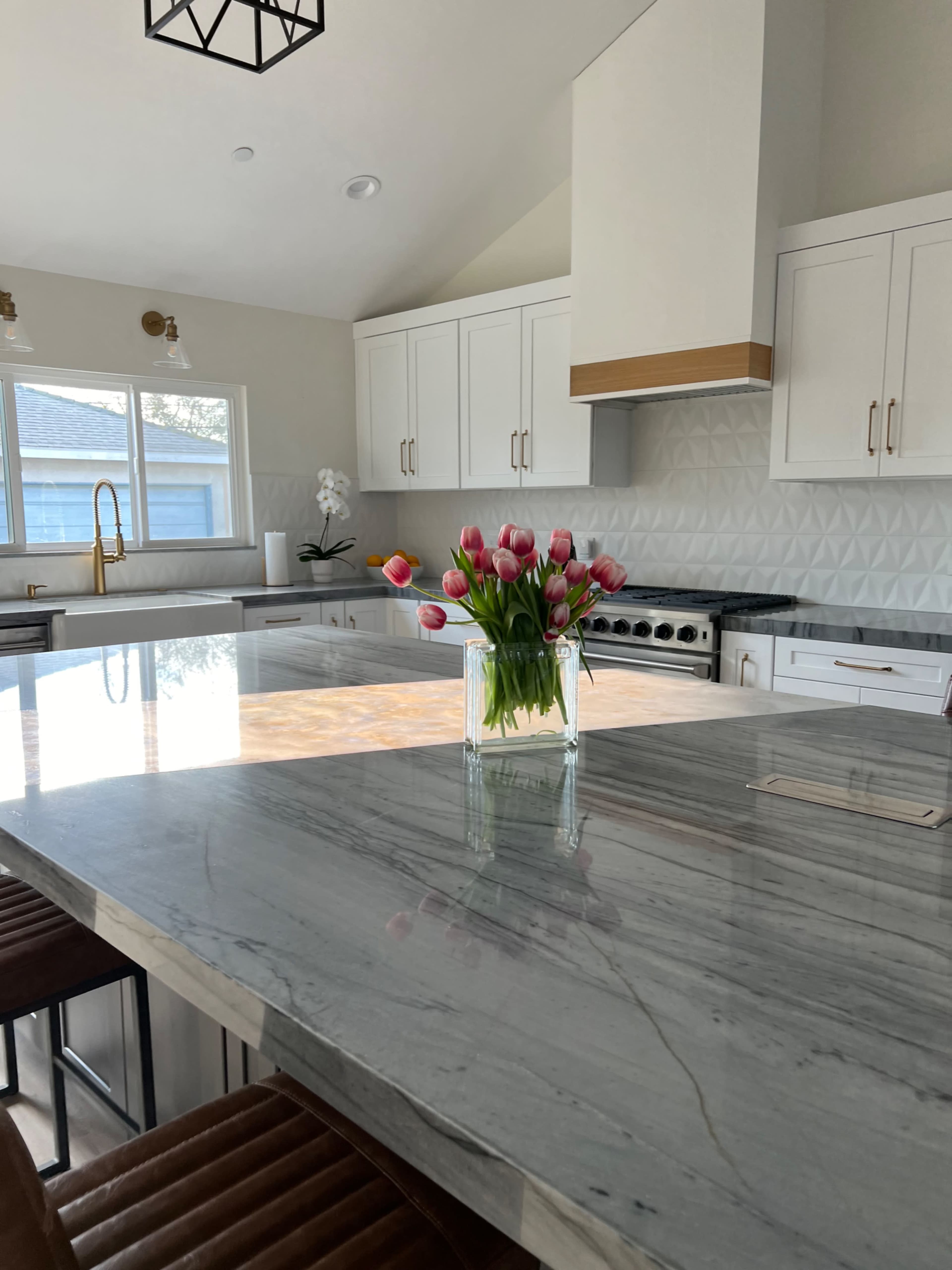 A modern kitchen features a large marble countertop with a vase of tulips at its center, surrounded by bar stools and sleek cabinetry.