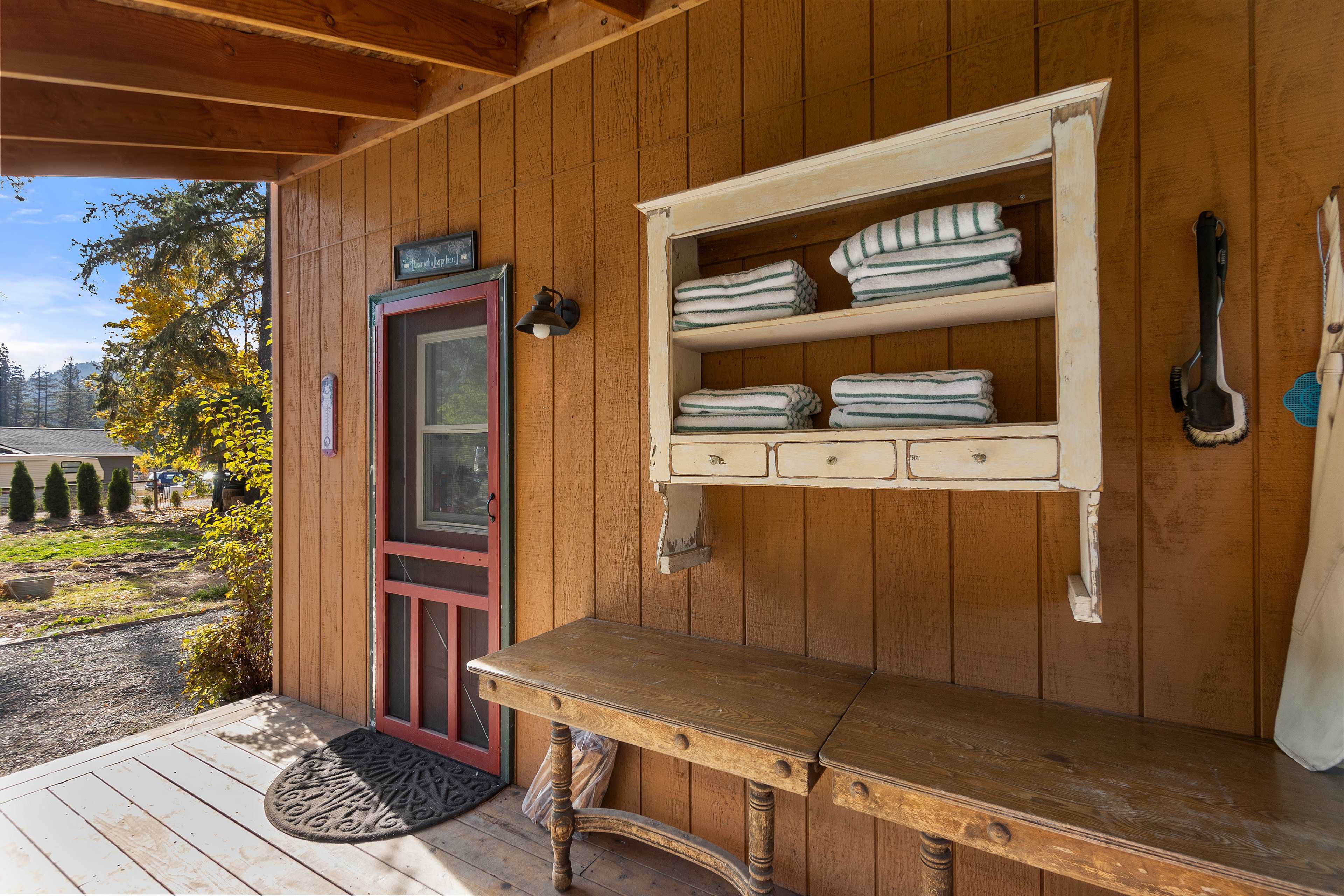 The image shows a wooden porch with a rustic table and a shelf holding neatly folded towels, alongside a red door framed by wooden paneling.