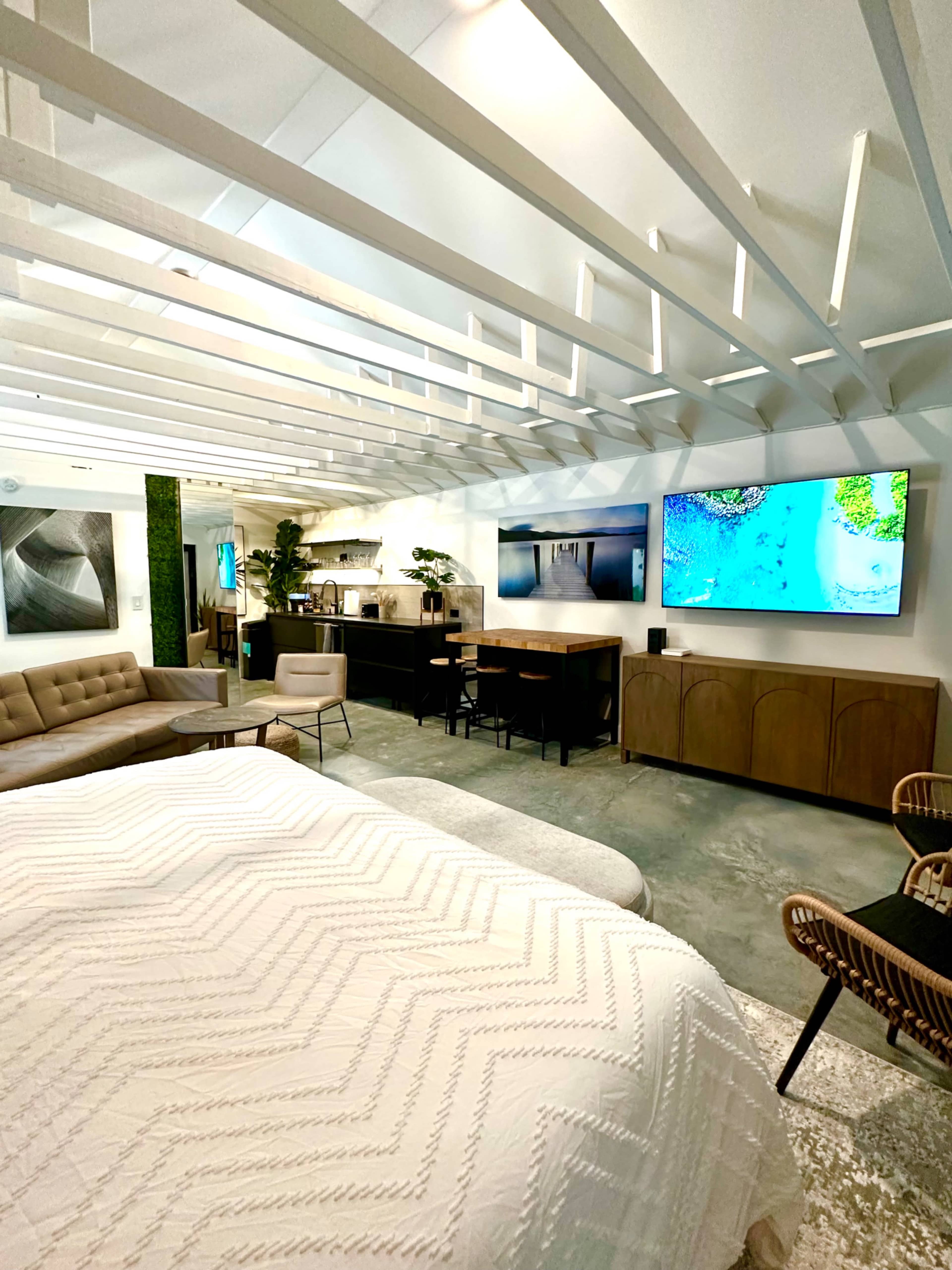 A modern living space featuring a comfortable bed, a sitting area with a sofa and chairs, and a kitchen area with two mounted televisions.