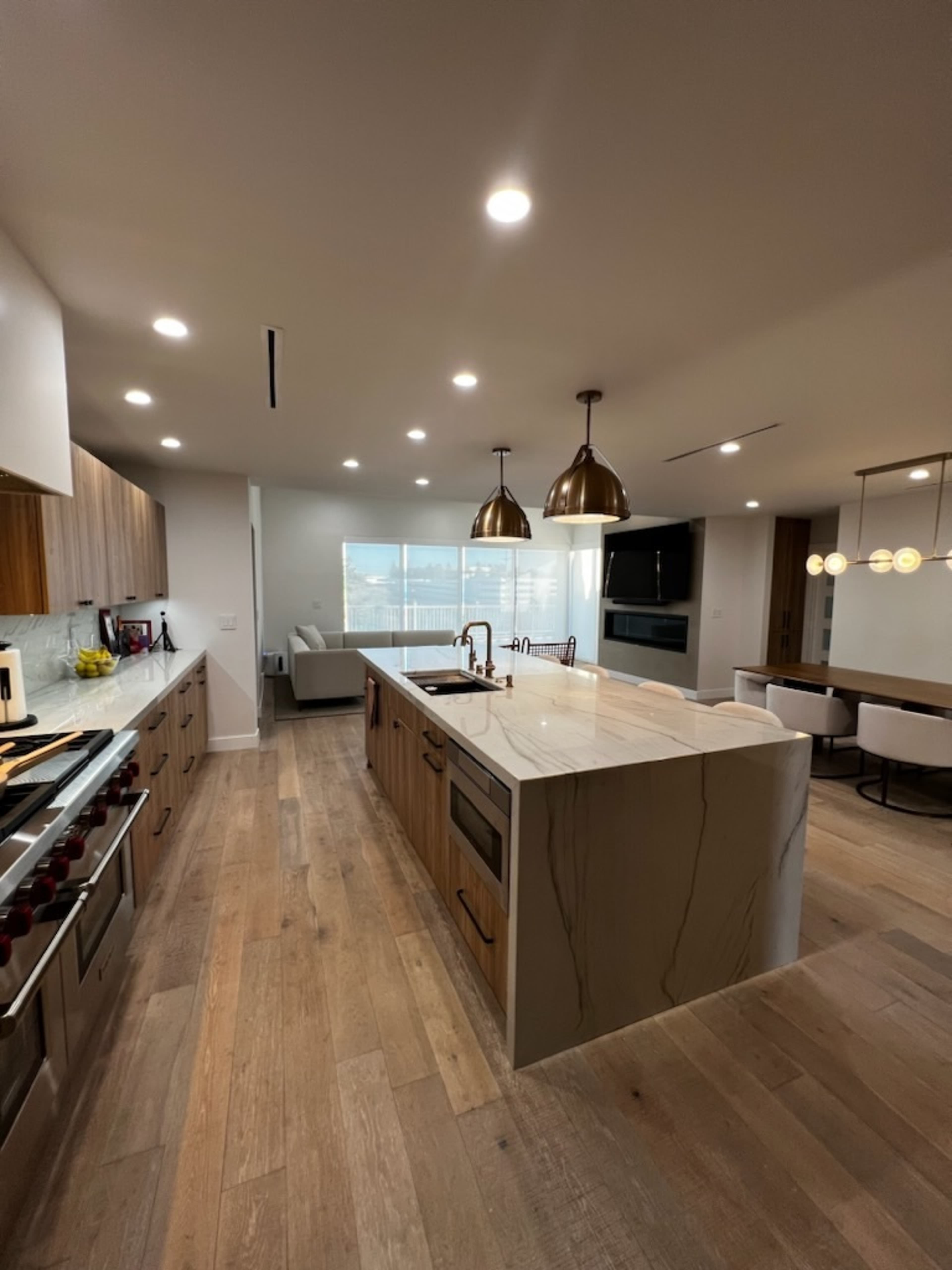 The kitchen features an island with a marble countertop, stainless steel appliances, and pendant lighting, leading to a large window with a view of the outside.