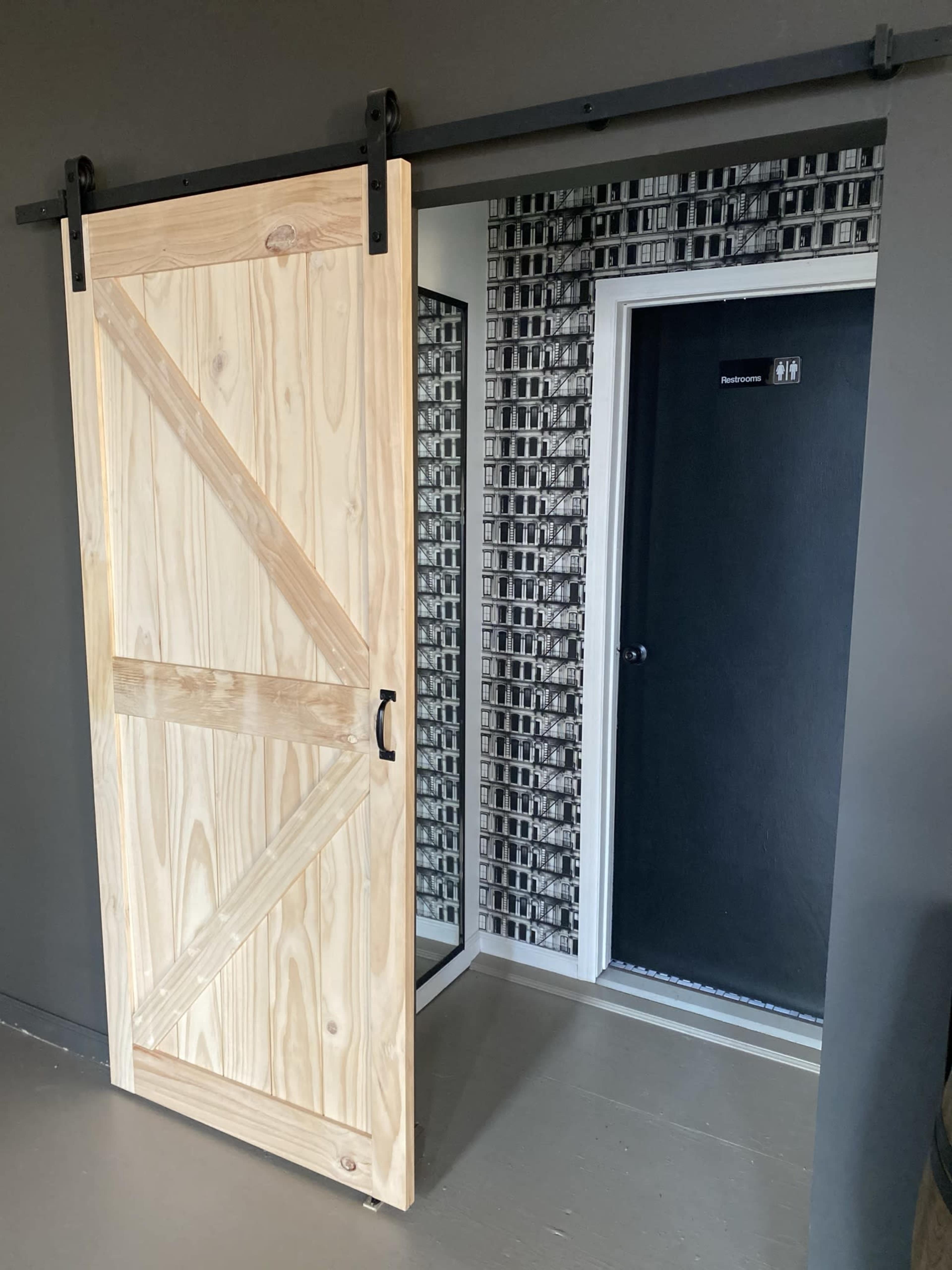 A sliding wooden barn door partially open, revealing a dark door labeled with a restroom sign, situated next to a wall with a patterned black and white design.