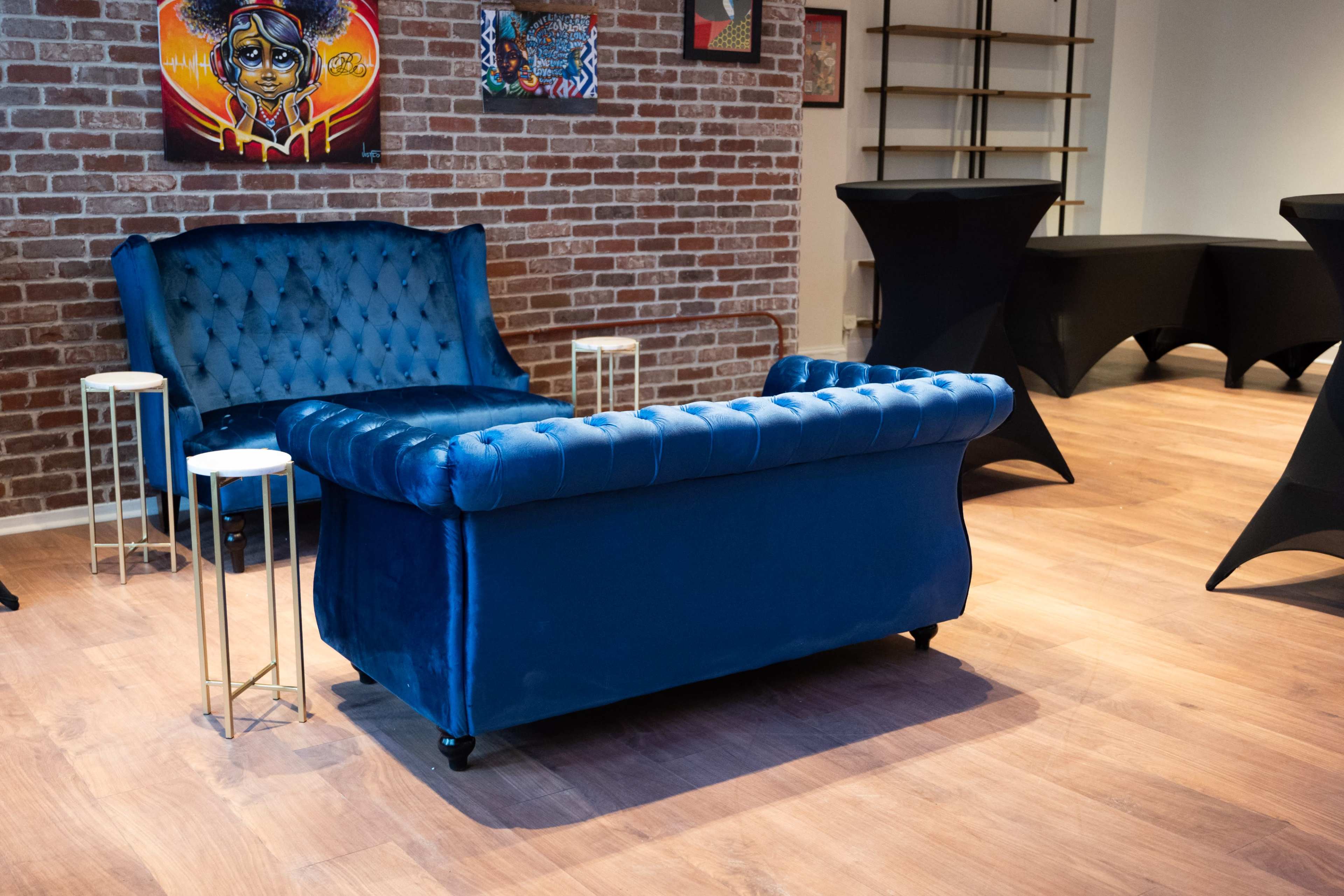 The image shows a blue velvet sofa in a room with exposed brick walls and small tables beside it.
