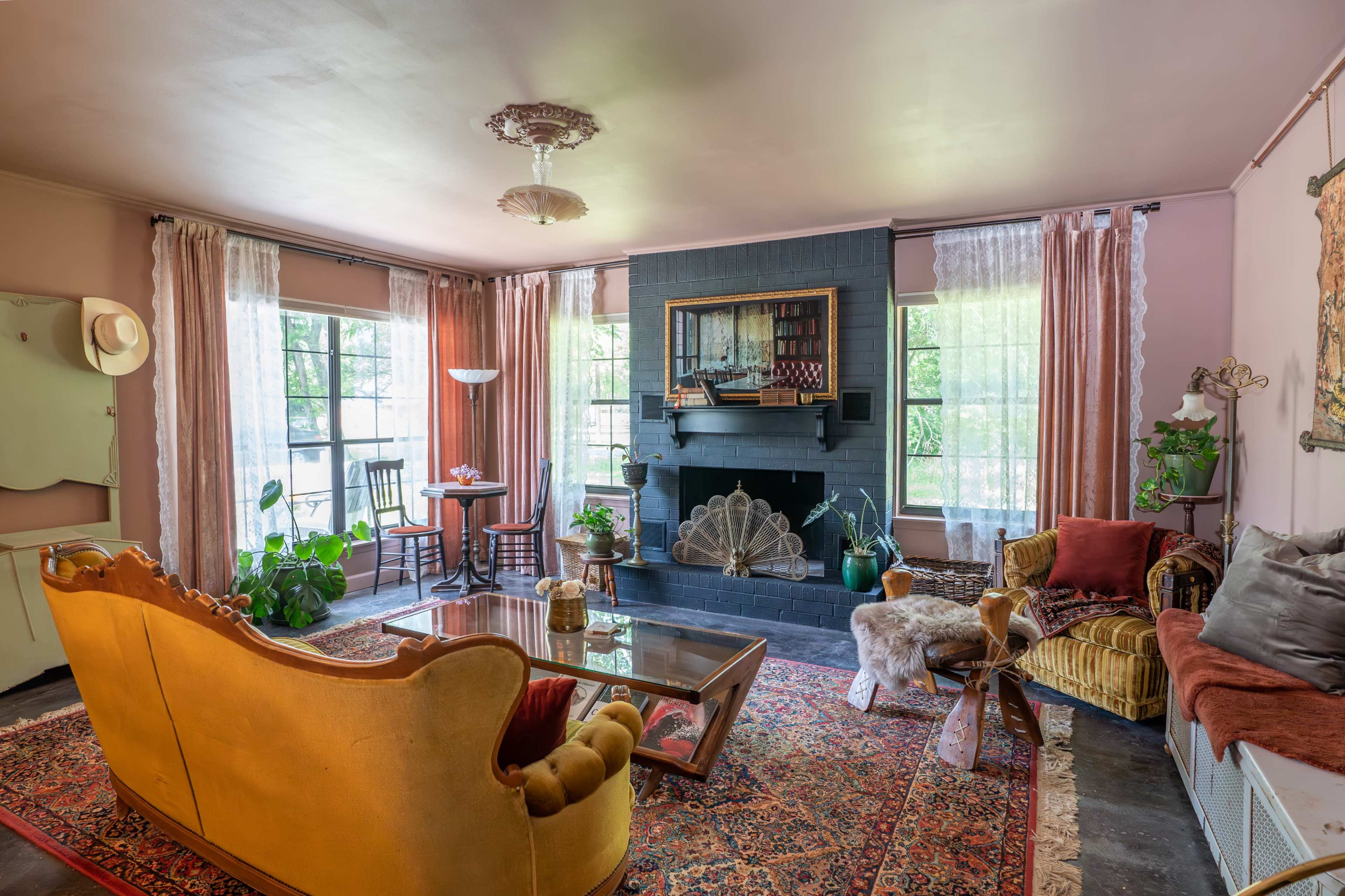 The living room features a mix of vintage furniture, a fireplace with decorative accents, and large windows allowing natural light to fill the space.