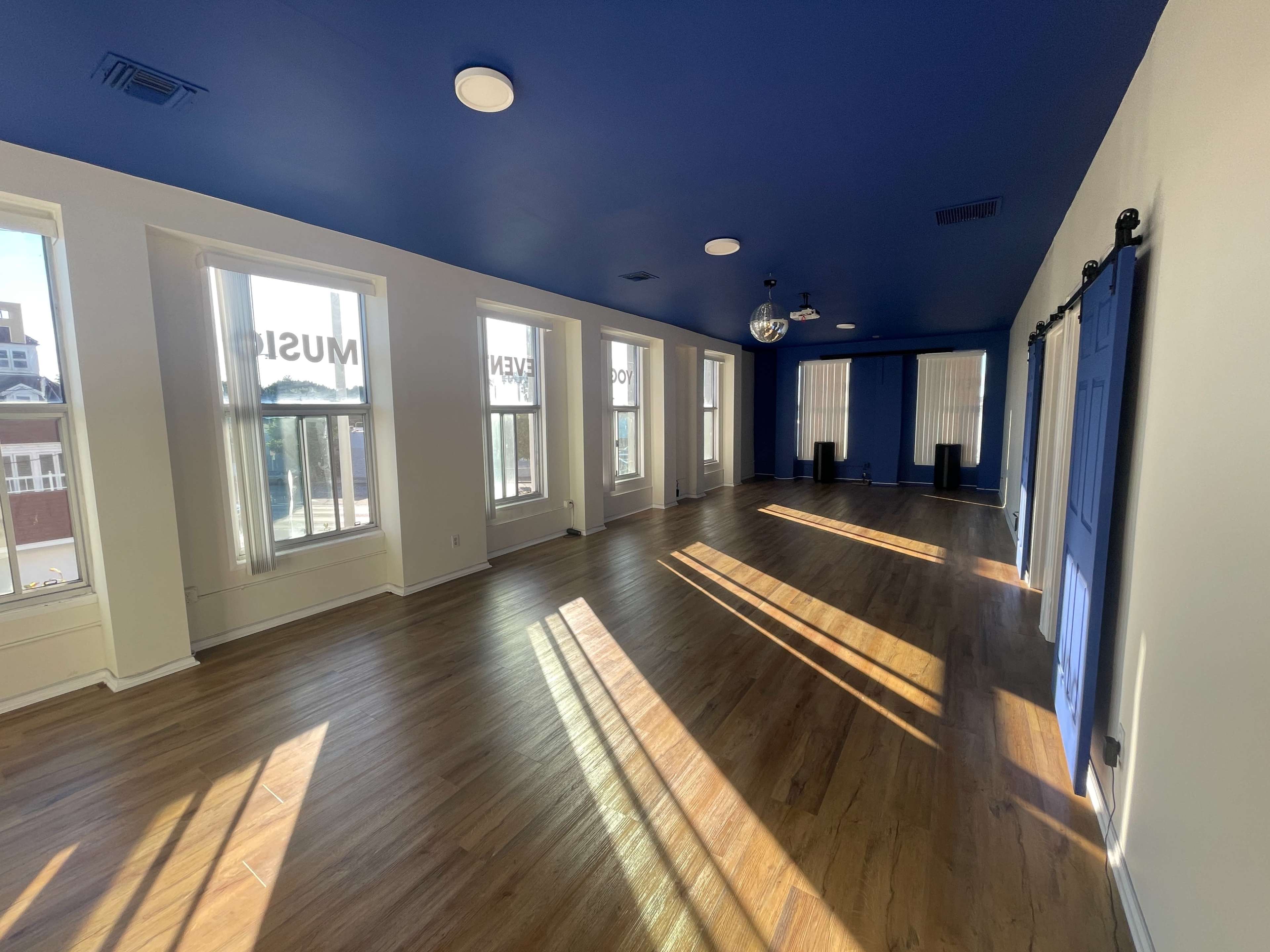 The image shows a spacious, brightly lit room with large windows, a blue ceiling, and wooden flooring, featuring a disco ball and two columns of light streaming through the windows.