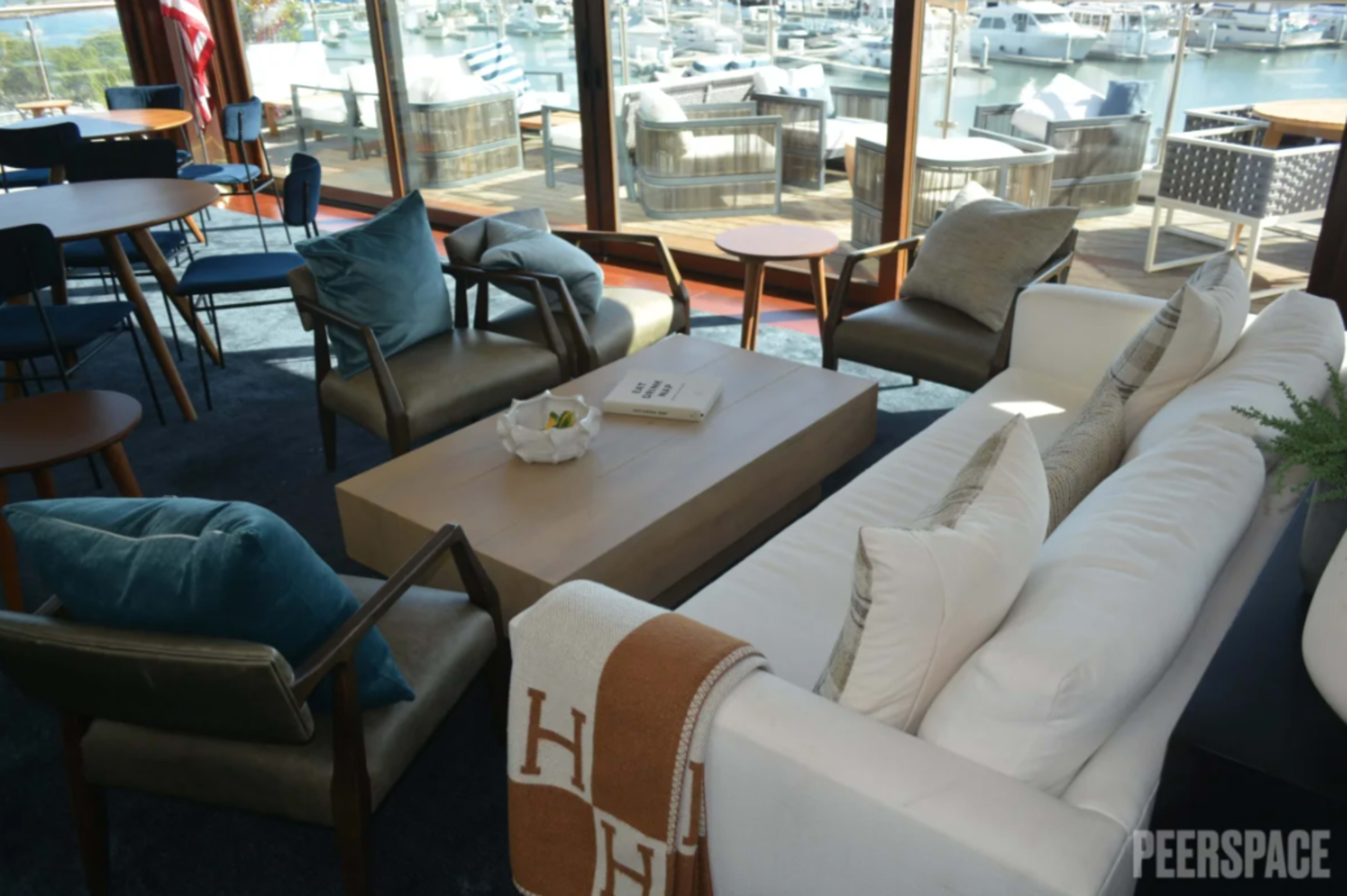 The image shows a modern living space with a white sofa, a wooden coffee table, and several chairs, overlooking a marina filled with boats.